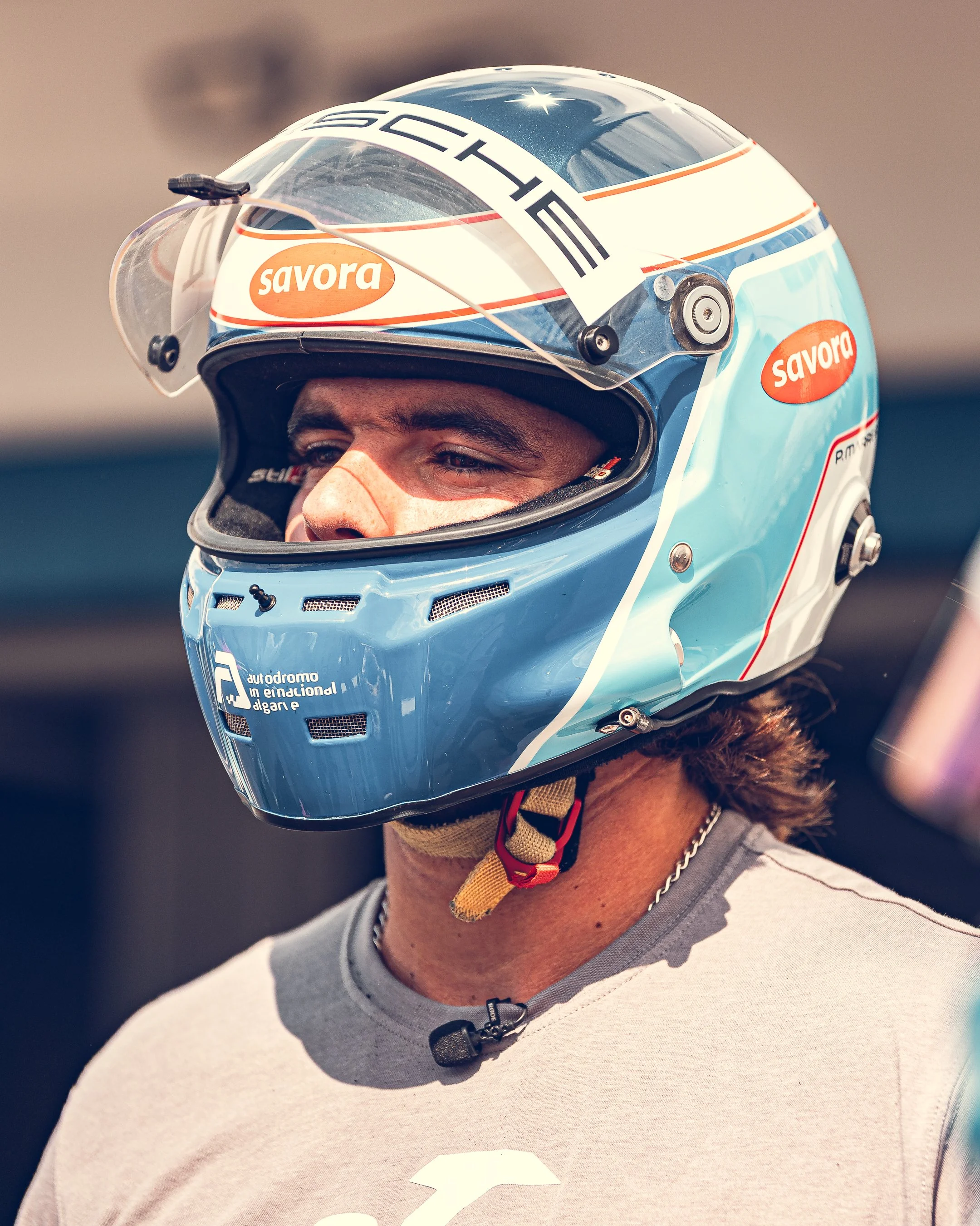 Close-up of a person wearing a blue racing helmet with sponsorship logos, including 'Savora' and 'F'. The helmet has a clear visor and a yellow strap. The individual is dressed in a gray shirt with a black zipper and a chain necklace.