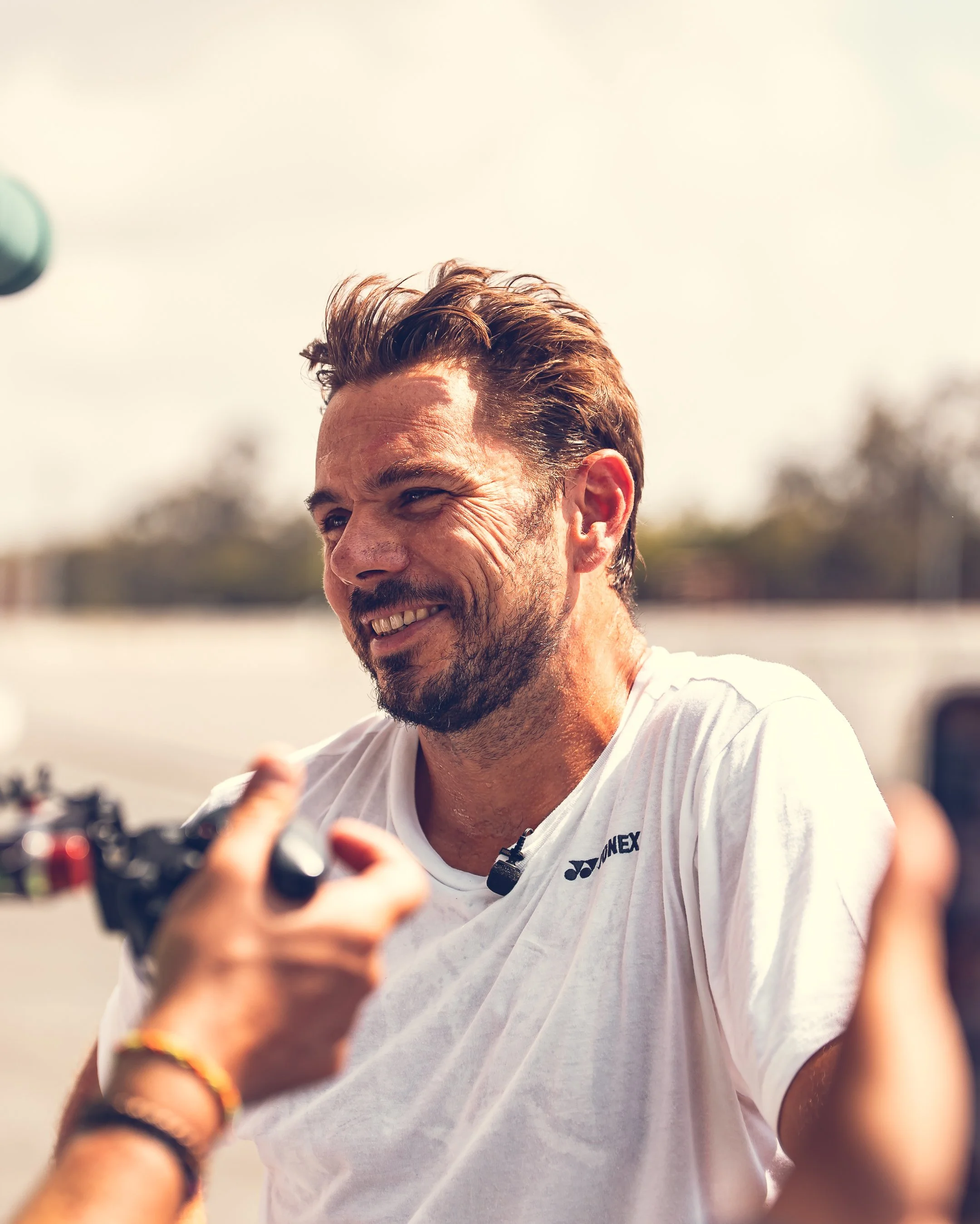 A man with dark brown hair and a beard smiling outdoors, holding a camera, wearing a white t-shirt.