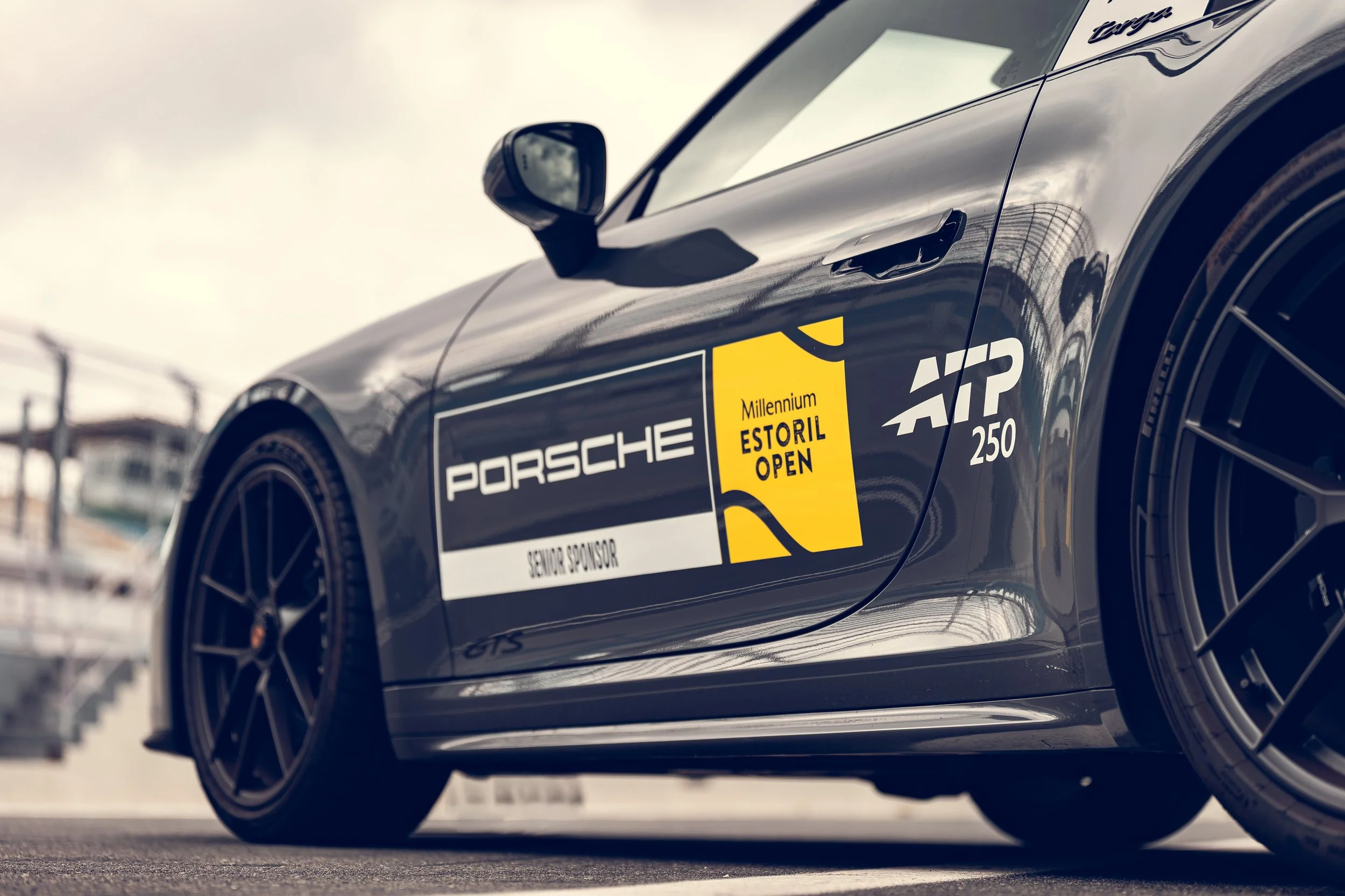 Close-up of a black Porsche race car with a yellow and black 'Millennium Estoril Open' sticker, parked on a racetrack with barbed wire fences in the background.