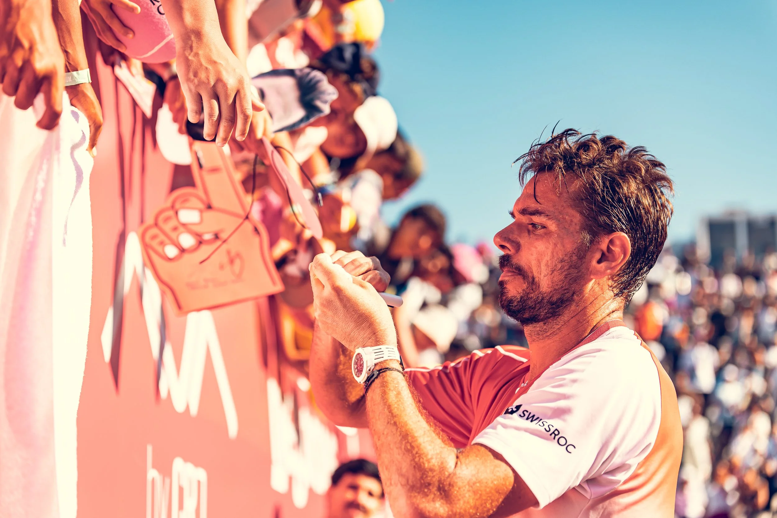 A man in a white and red sports shirt signing an autograph on a pink board during an outdoor sports event.