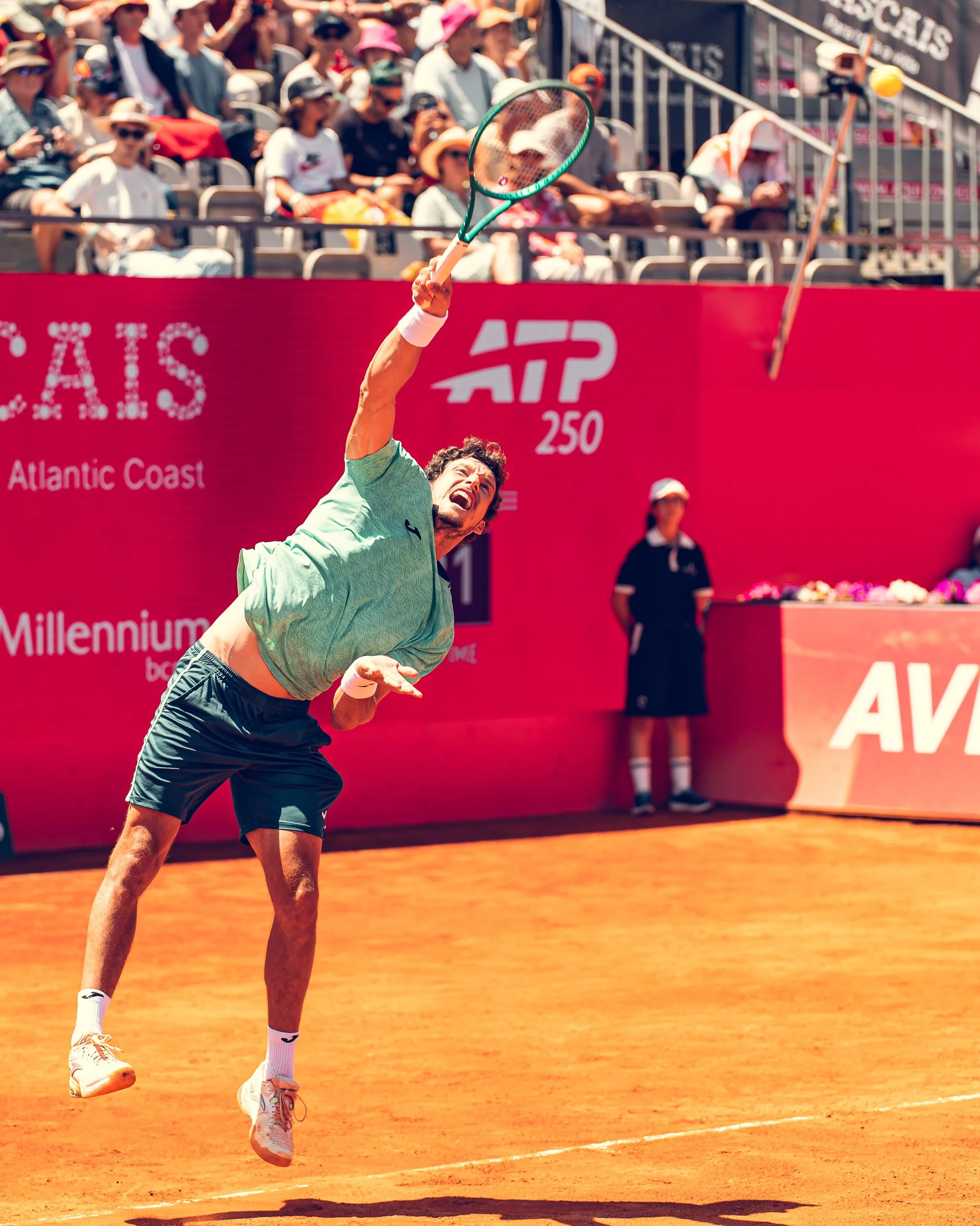 A male tennis player with curly hair, wearing a green shirt, black shorts, white socks, and sneakers, hits a tennis ball with a racket on a clay court during a match at the ATP 250 tournament at Atlantic Coast.