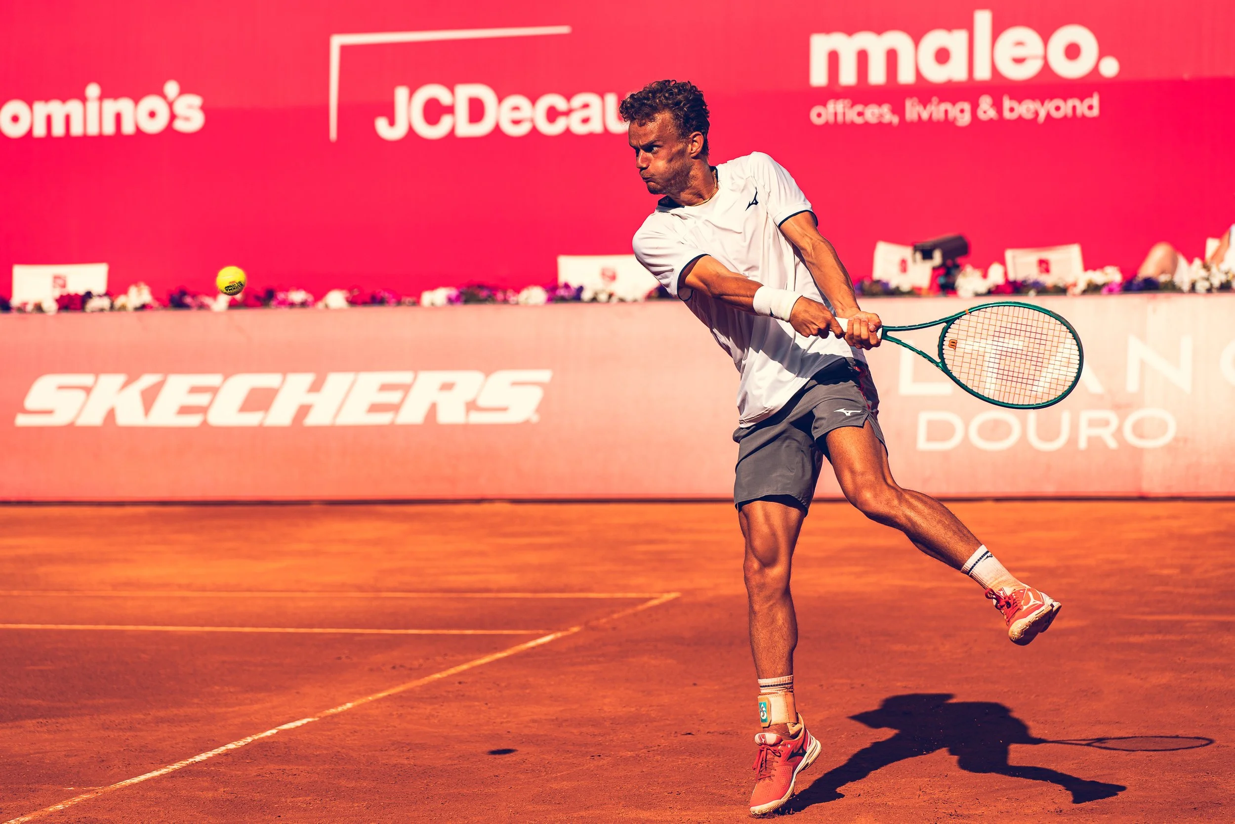 A man playing tennis on a clay court, wearing a white shirt, gray shorts, and red tennis shoes, hitting a yellow tennis ball.