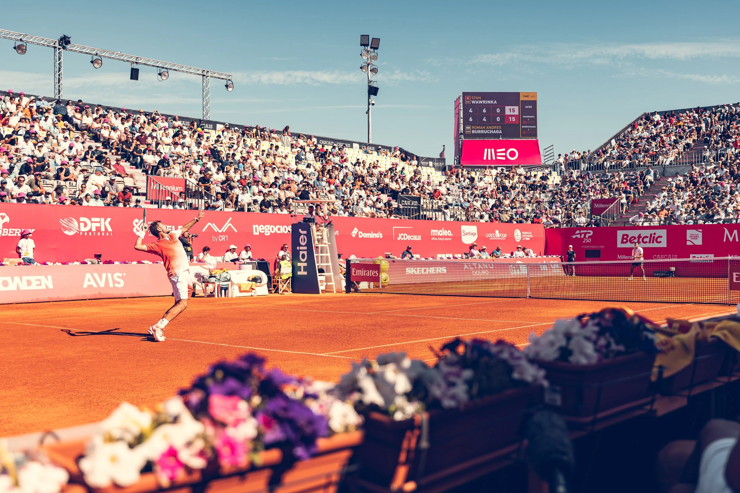 A tennis player in a red shirt and white shorts on a clay court during a match, with a large audience in the stands and a scoreboard showing a close score, surrounded by advertisements and colorful flowers in the foreground.