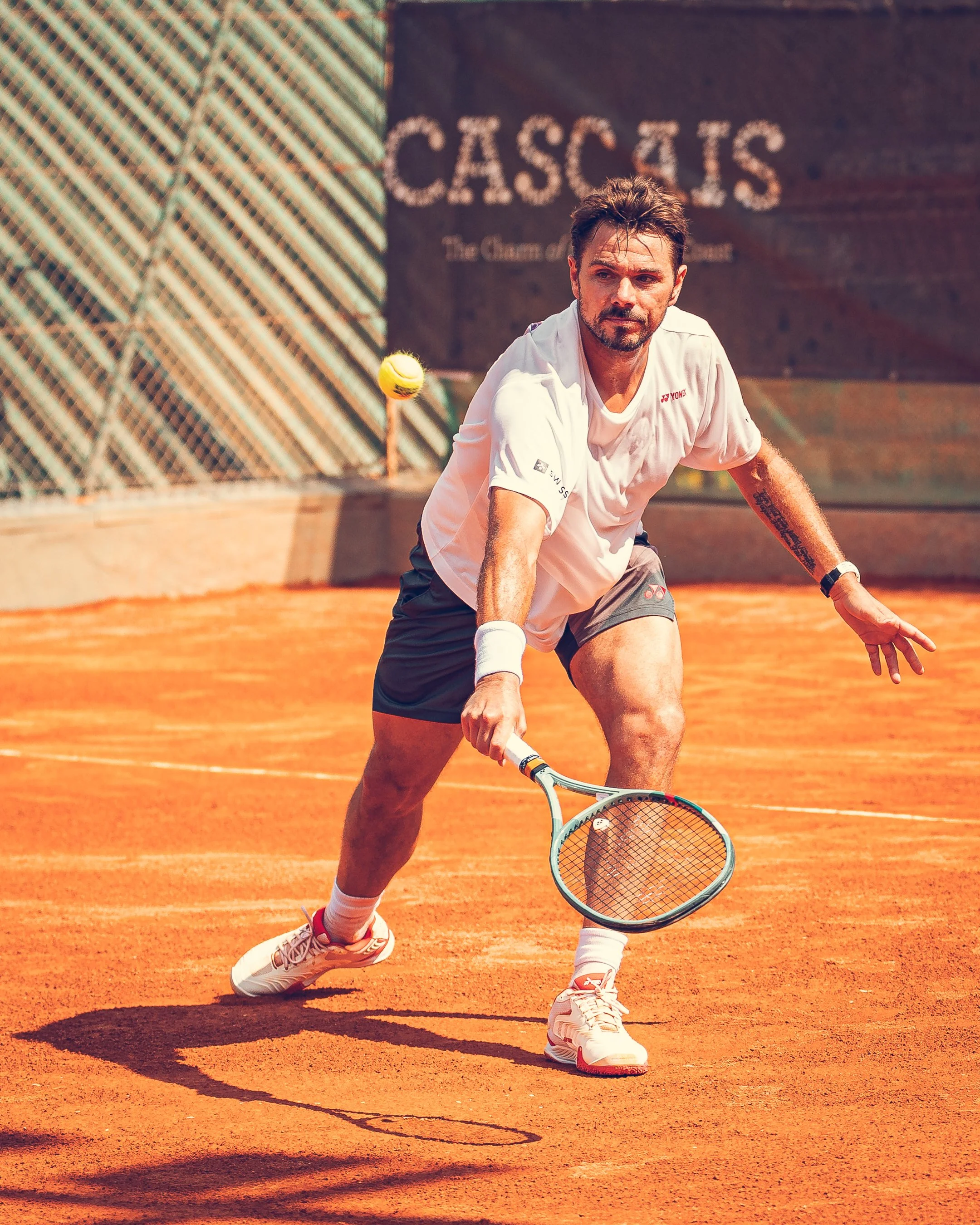 A tennis player in white shorts and a white T-shirt preparing to hit a tennis ball on a clay court, with a wooden barrier and a sign reading 'CASCAS' in the background.