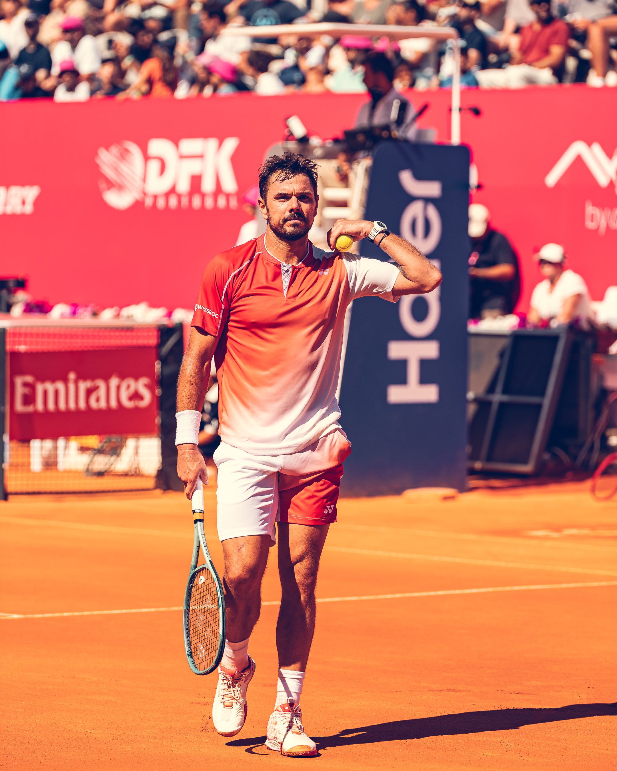 A male tennis player walking on a clay tennis court holding a tennis ball and a tennis racket, with a crowd of spectators in the background.