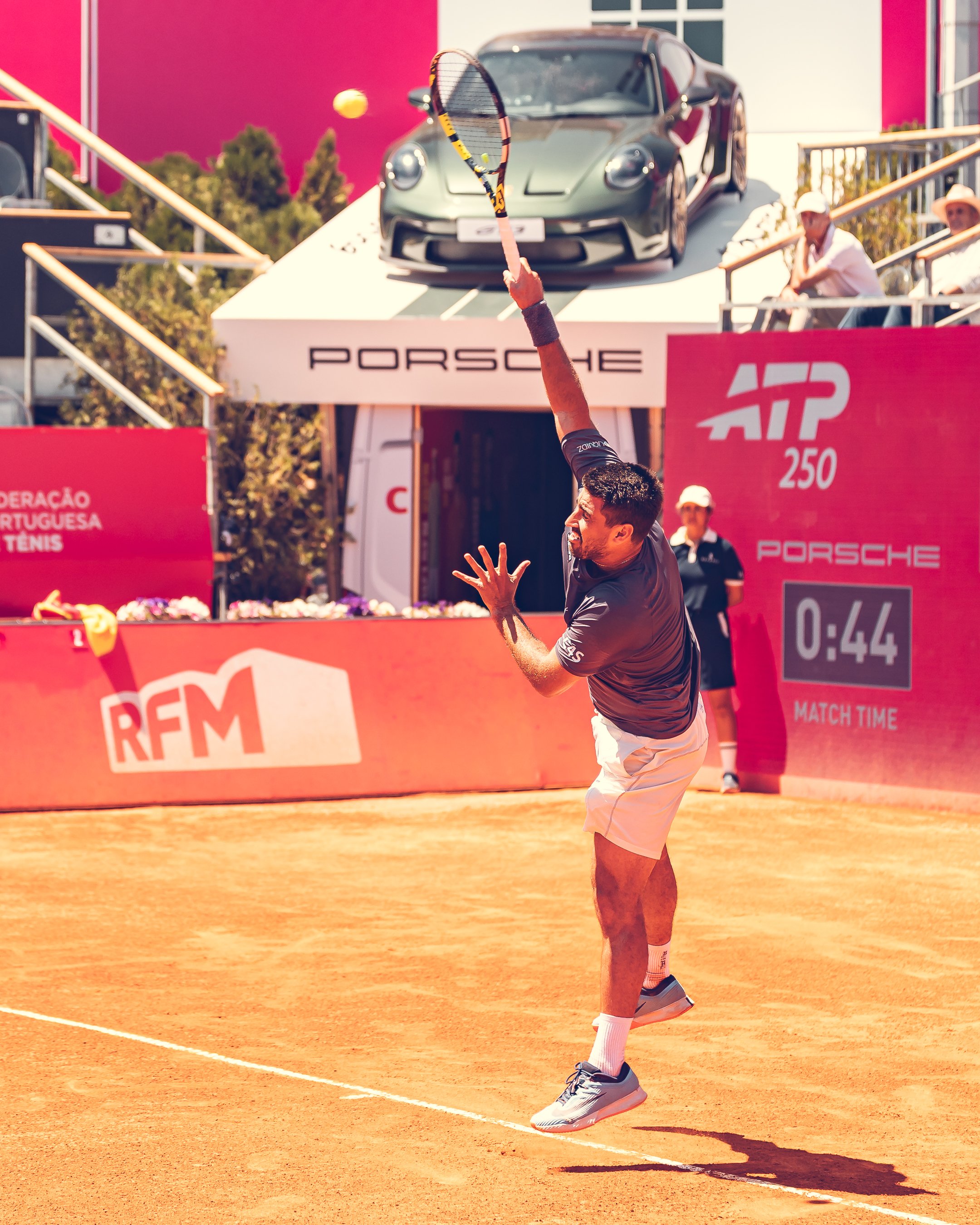 A man playing tennis on a clay court during daytime, with a black sports car on an elevated platform in the background and spectators sitting nearby.