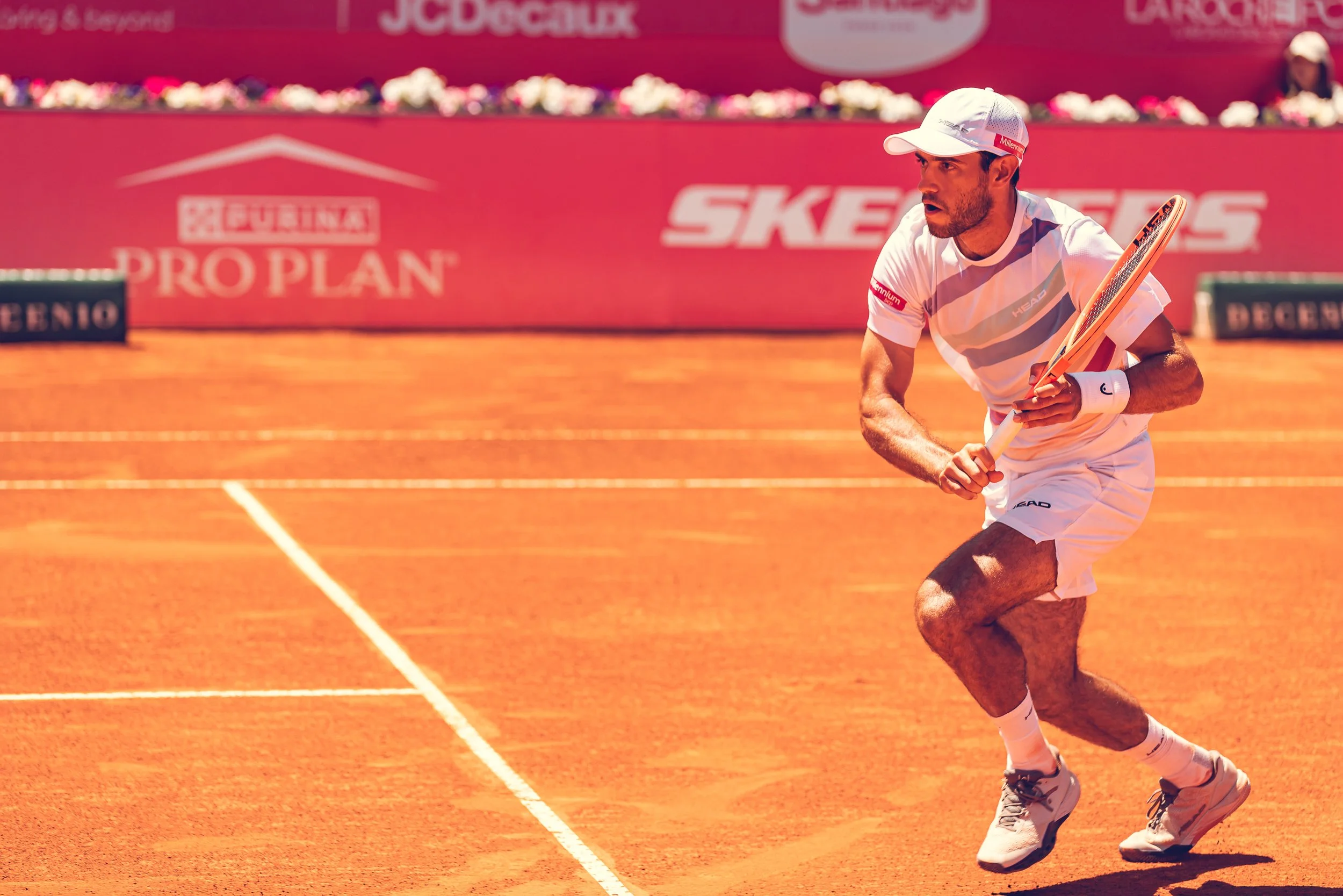 A male tennis player in a white uniform and cap is running on a red clay court during a match, holding a tennis racket. The background features a pink banner with sponsorship logos and a person wearing a cap sitting in the stands.