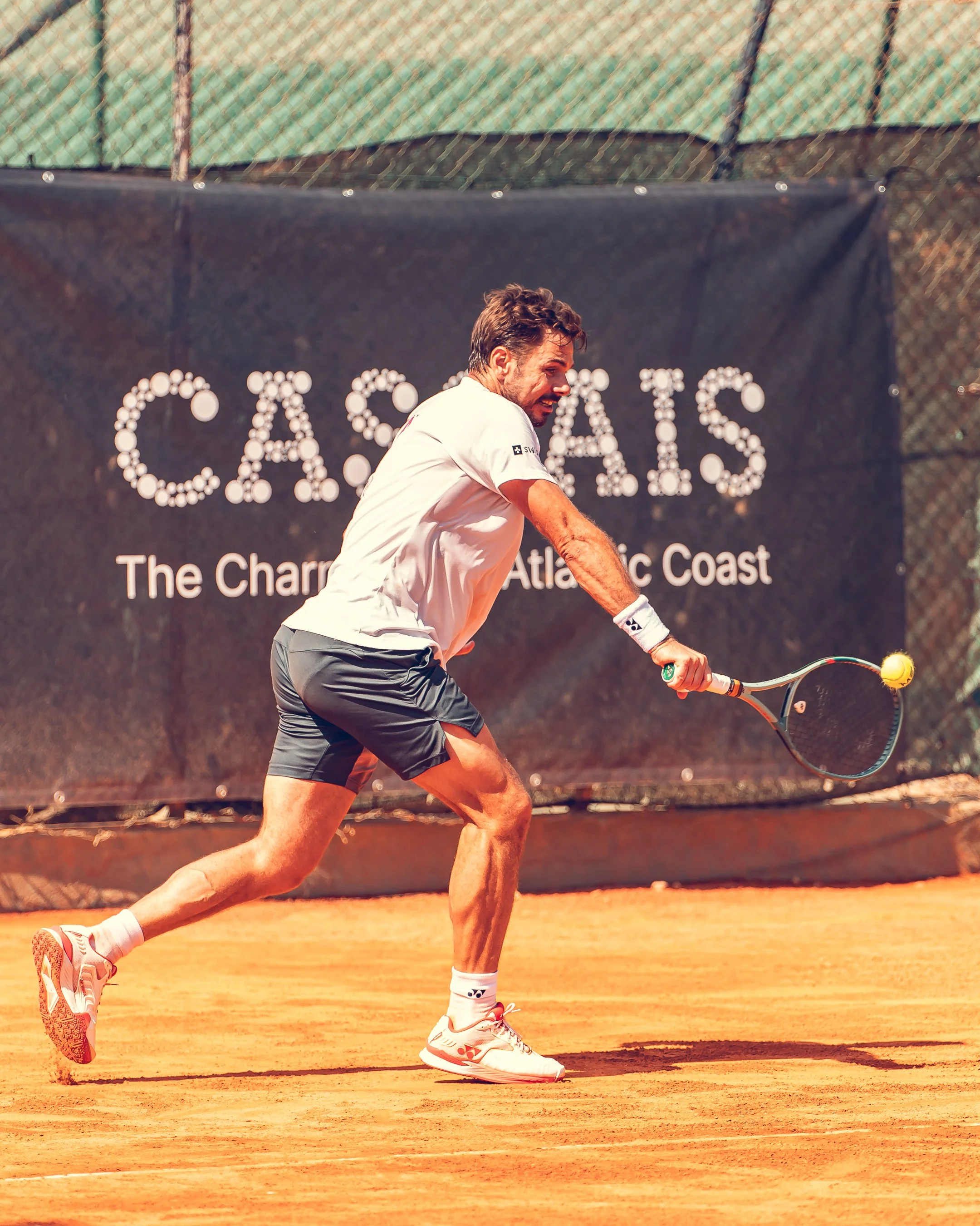 Male tennis player hitting a tennis ball on clay court, wearing white sportswear and white sneakers, with a chain-link fence and promotional banner in the background.