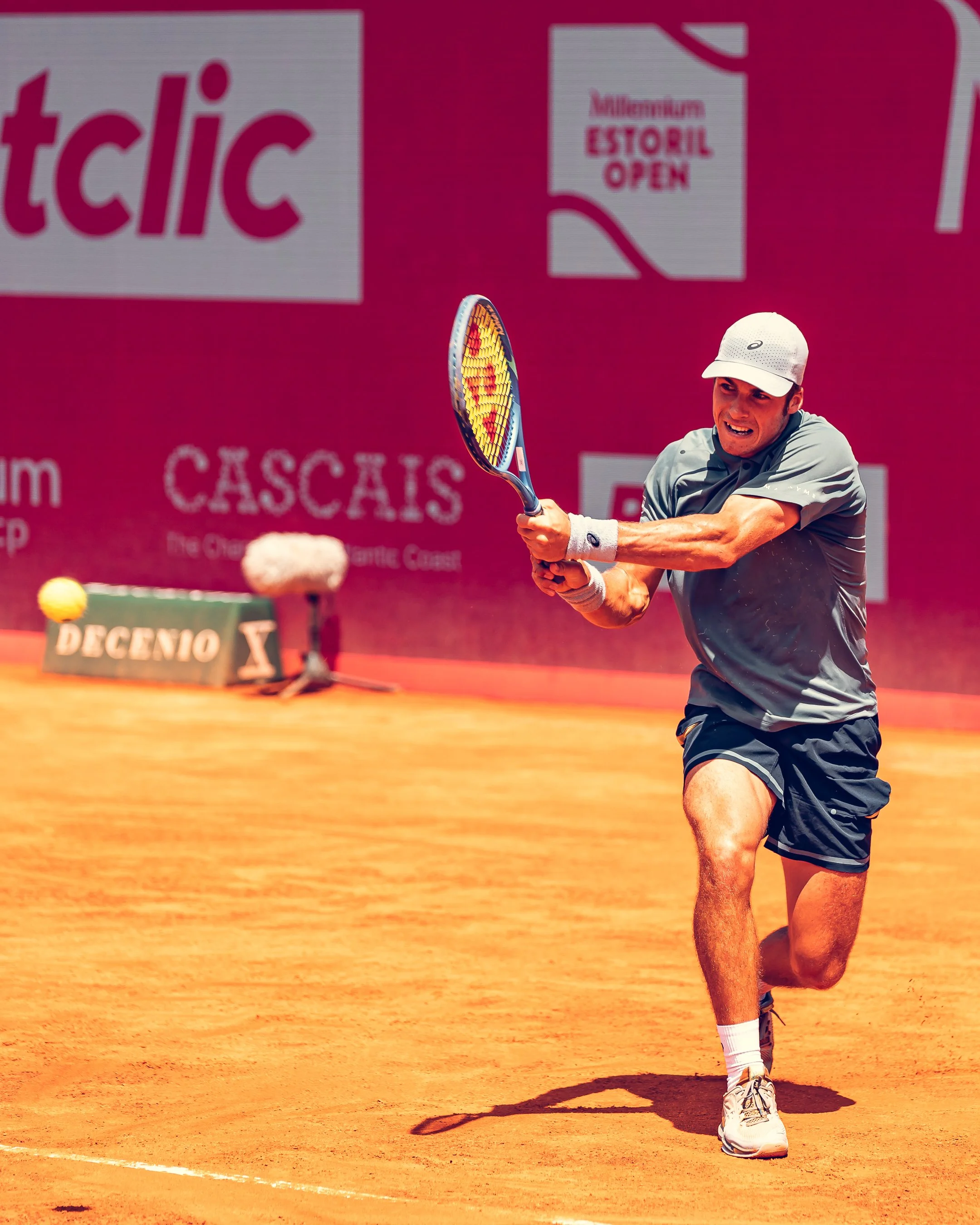 A male tennis player in athletic clothing and white cap hitting the ball on a clay court during a match.
