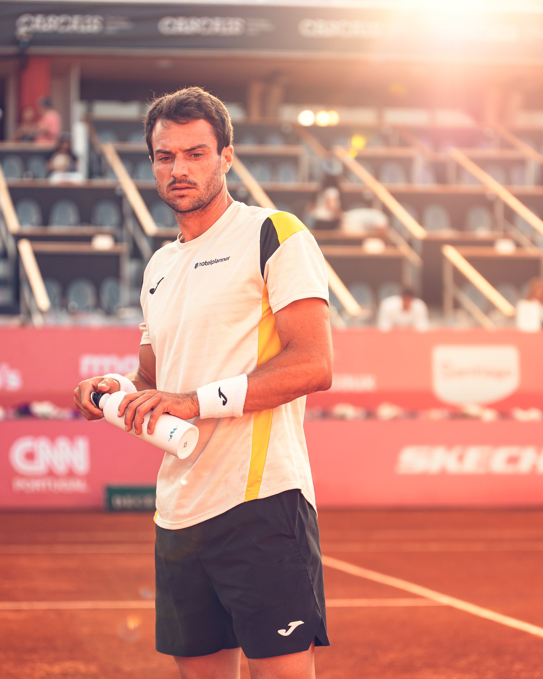 A male tennis player holding a water bottle on a tennis court during daylight sunset, wearing a white and yellow sports shirt and black shorts with a logo, with blurred spectators in the background.