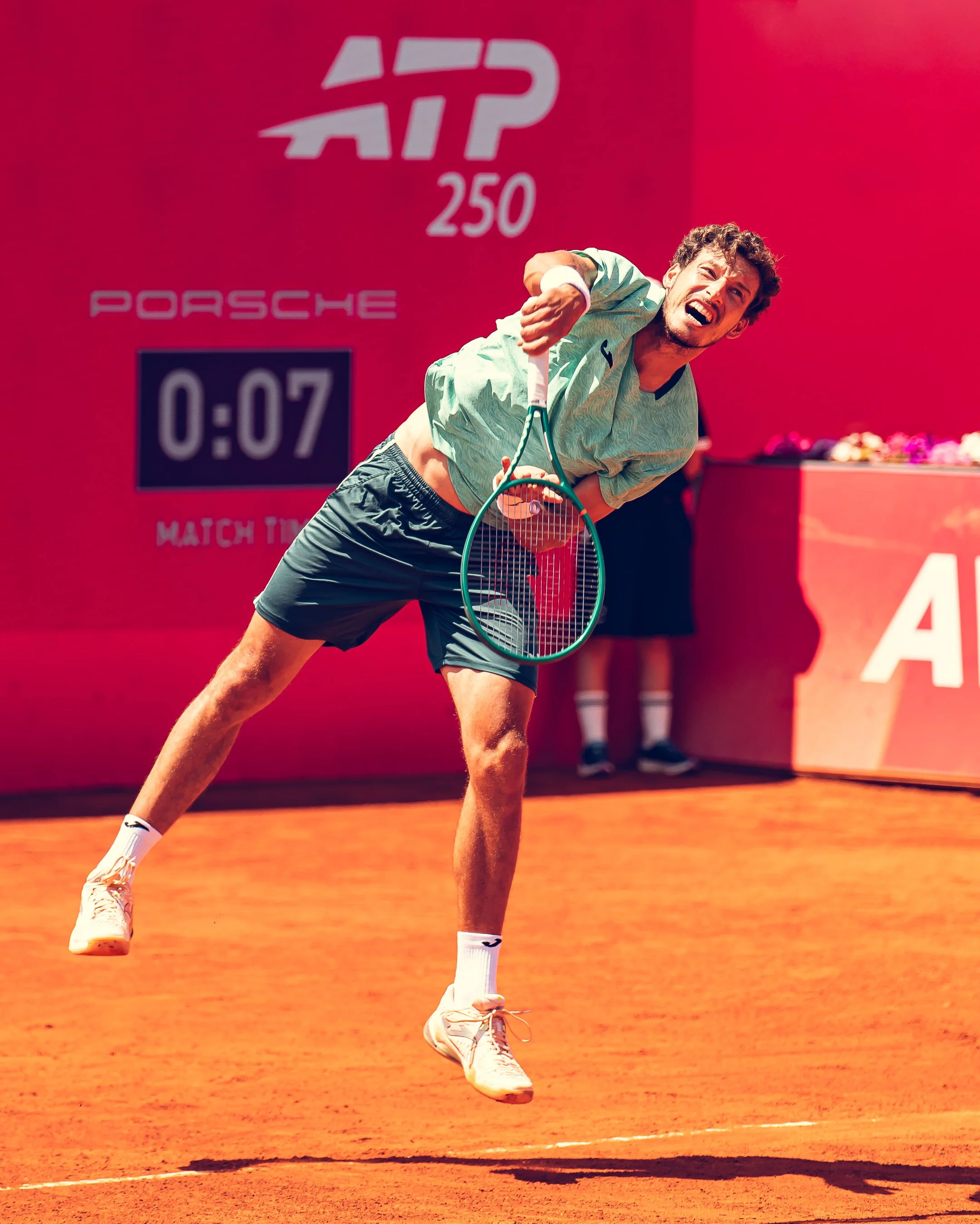 A man on a tennis court appears to be falling or tripping, holding a tennis racket, with a bright pink backdrop displaying the ATP 250 logo, a digital scoreboard showing 0:07, and a table with flowers in the background.