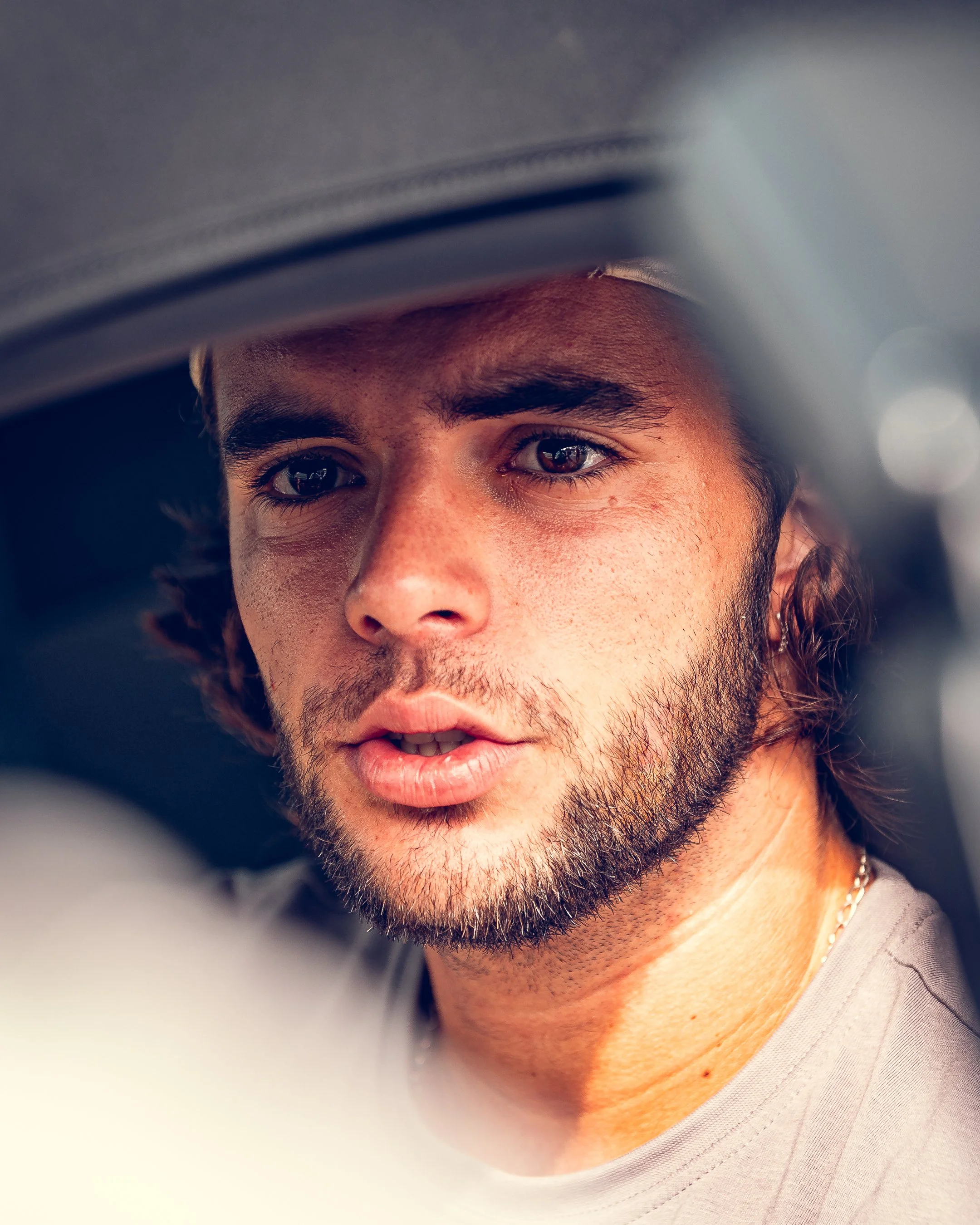 Close-up of a young man with dark hair, brown eyes, and a beard, wearing a light-colored shirt and a necklace, with a neutral background.