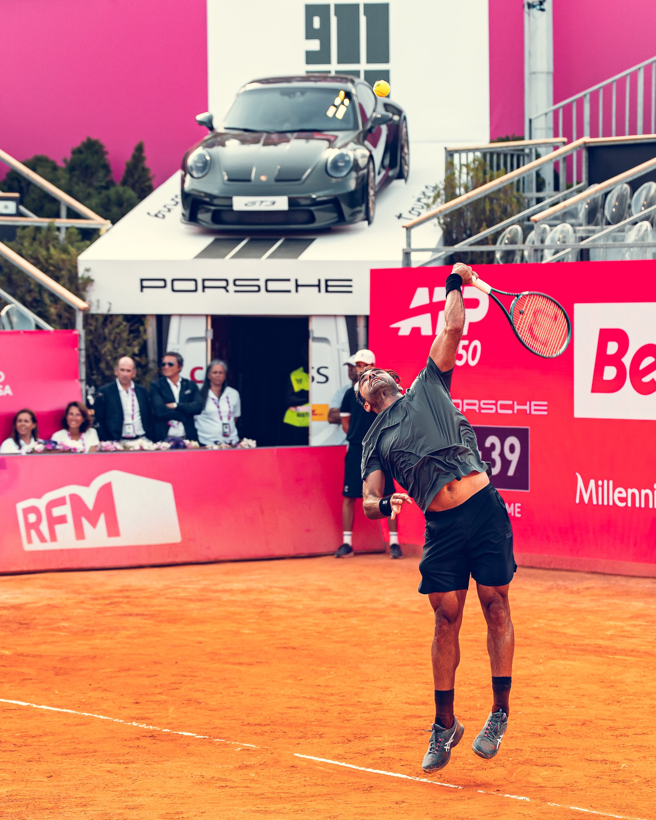A professional tennis player in mid-air hitting a shot on a clay court during a match, with a backdrop of spectators and a Porsche advertisement.