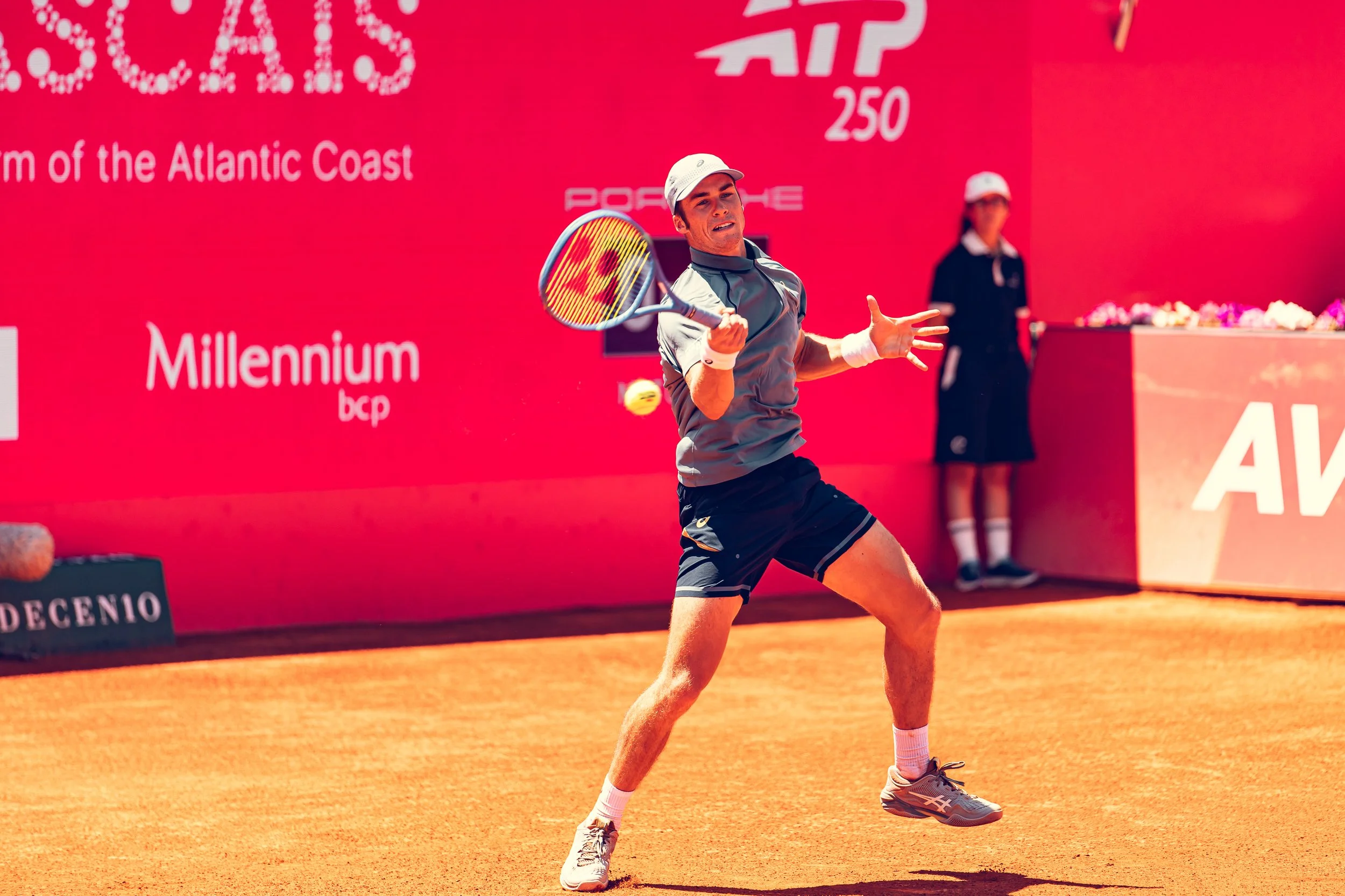 A male tennis player in action on a clay court during a match, with a red backdrop and event signage in the background.