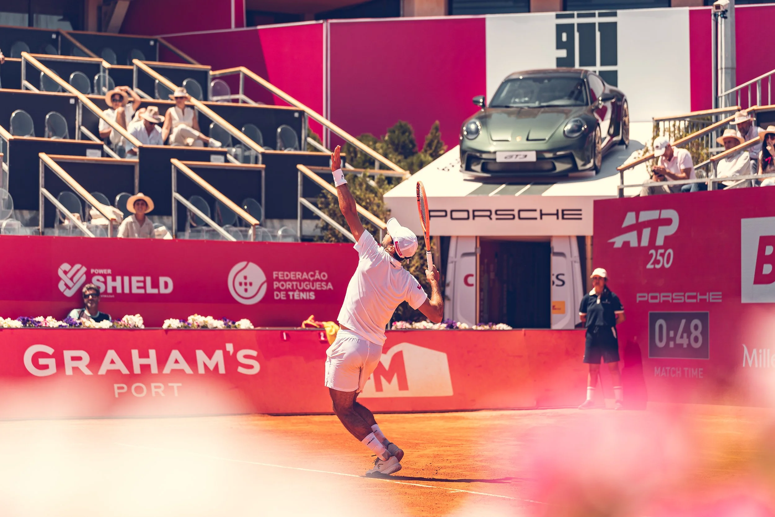 Tennis player in white outfit and cap playing on a clay court during a match. There are advertisement banners, including Graham's Port, and a black Porsche 911 car on a platform in the background. Spectators are seated in the stands, some wearing hat
