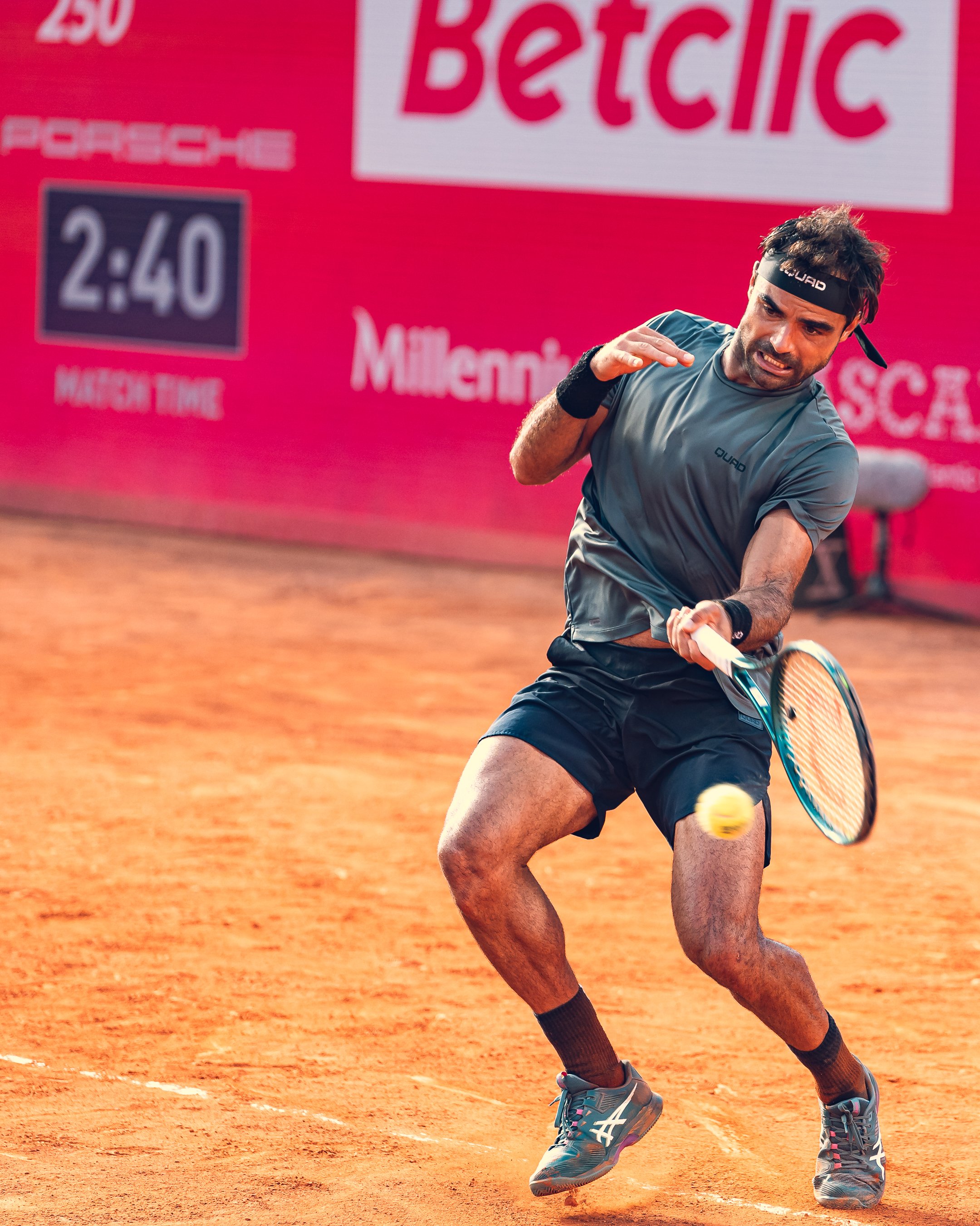 A male tennis player in a blue workout shirt and gray shorts hits a tennis ball on a clay court. The background shows red advertising banners, including the Betclic logo, and a digital scoreboard displaying 2:40.