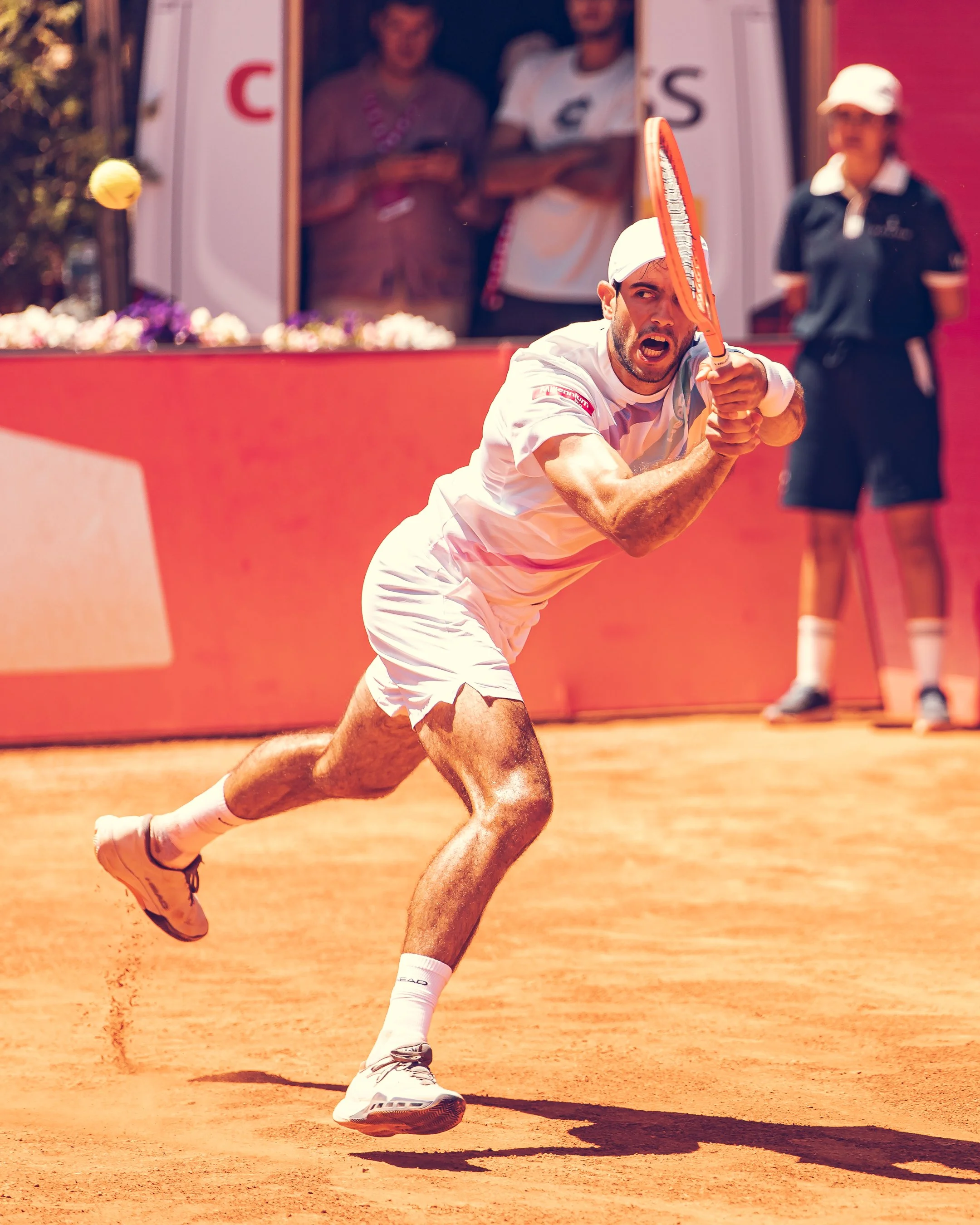 A tennis player in a white shirt and shorts is running on a clay court, holding a tennis racket with an intense expression, while a line judge in black uniform stands in the background.
