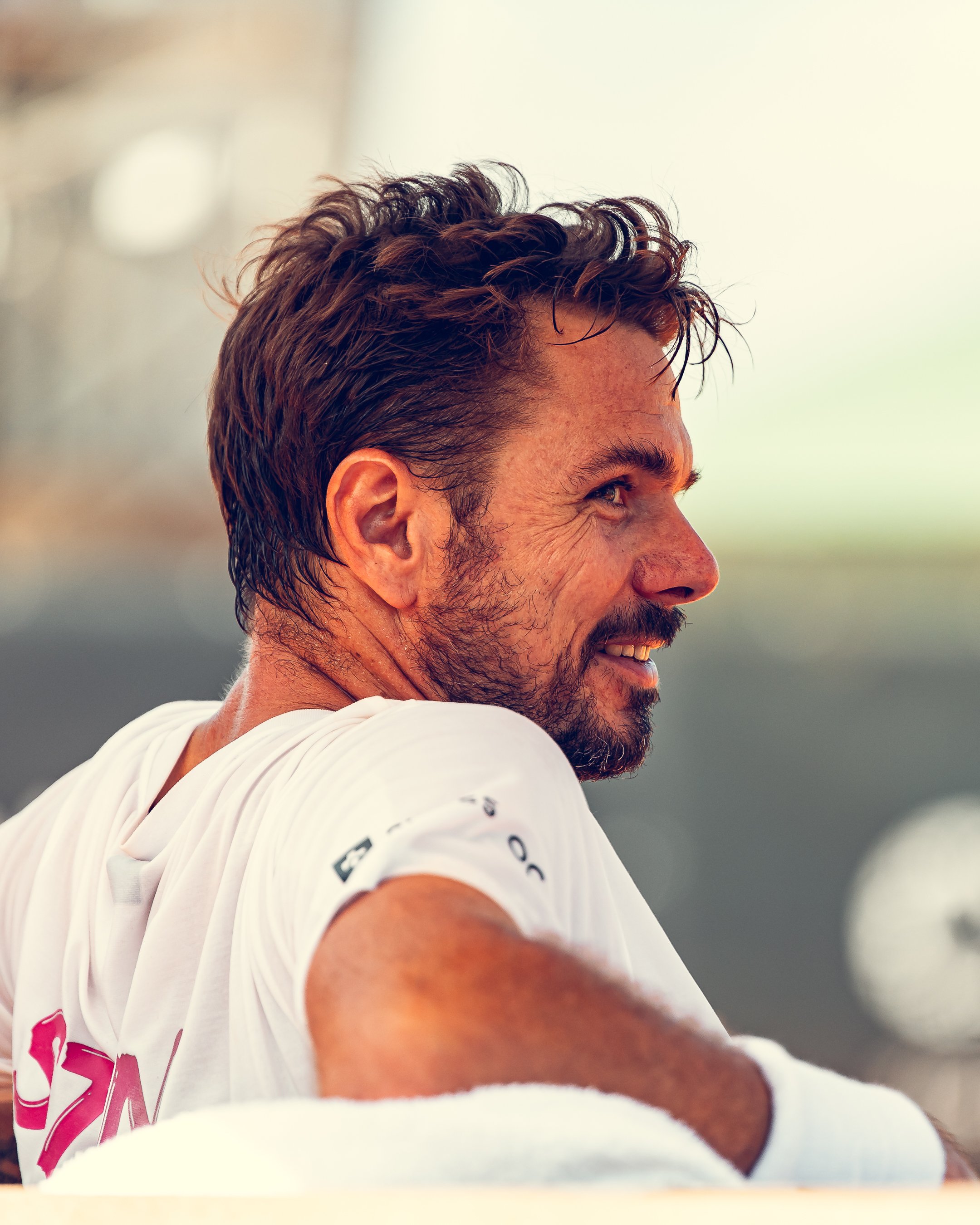 Close-up side profile of a man with messy brown hair and a beard, smiling, wearing a white t-shirt with red text on the back, outdoors during daytime.