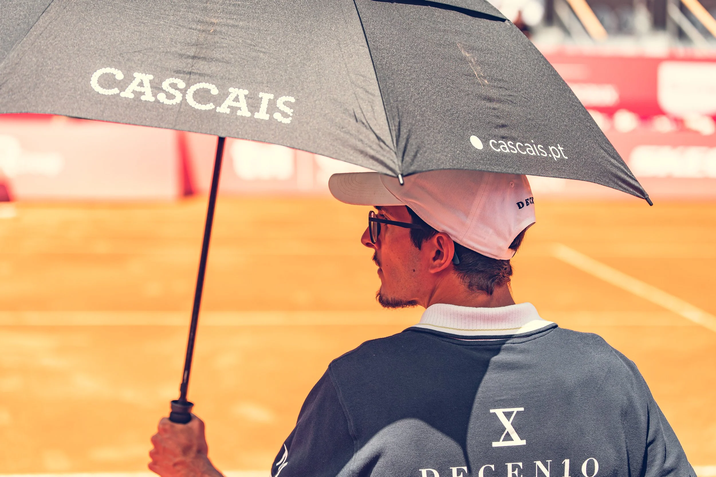 A man wearing glasses and a cap holding a black umbrella with the word 'Cascais' on it, standing on a tennis court.