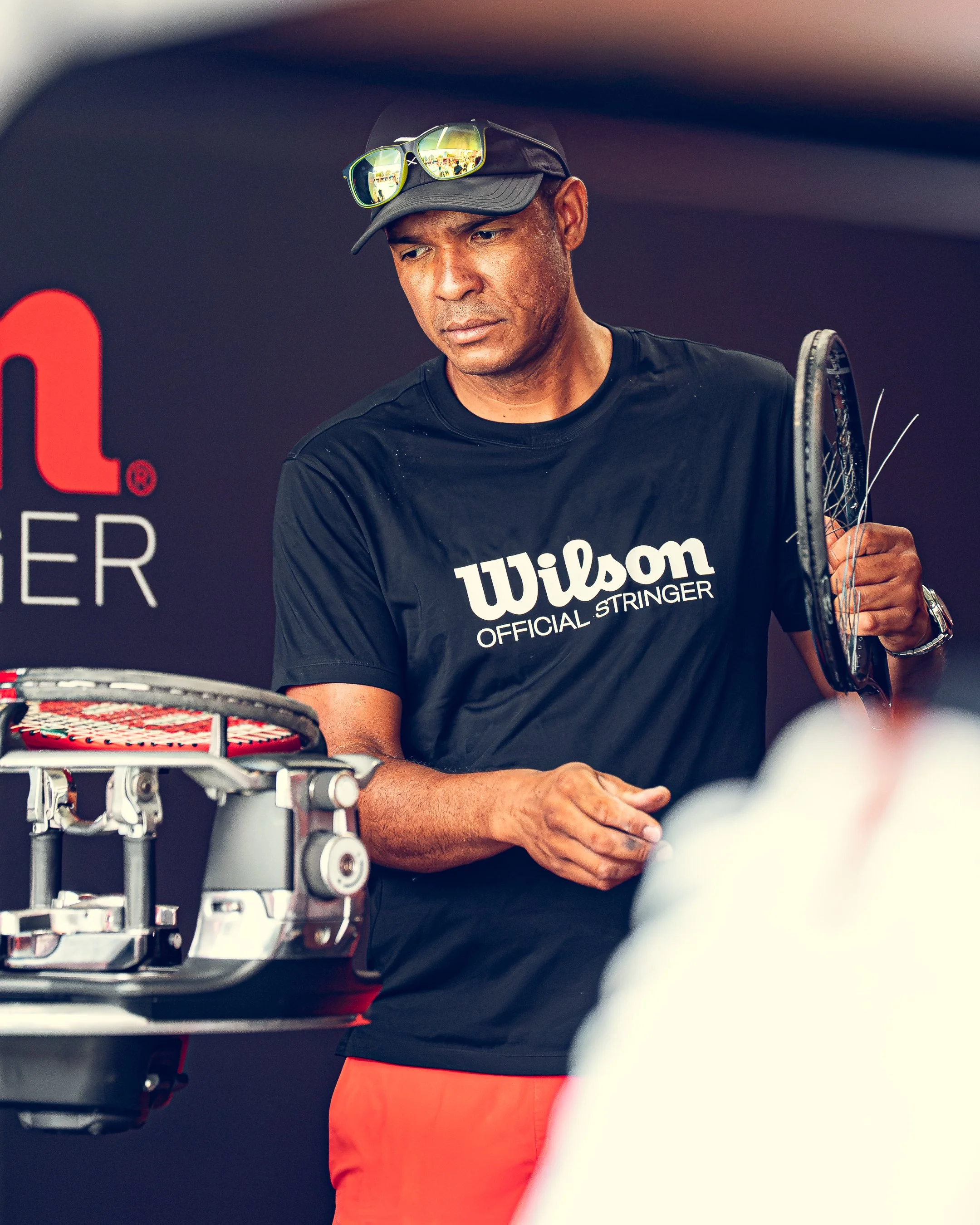 A man in black Wilson tennis apparel holding a tennis racket at a tennis match, with another person in white in the foreground and a black background with red and white text.