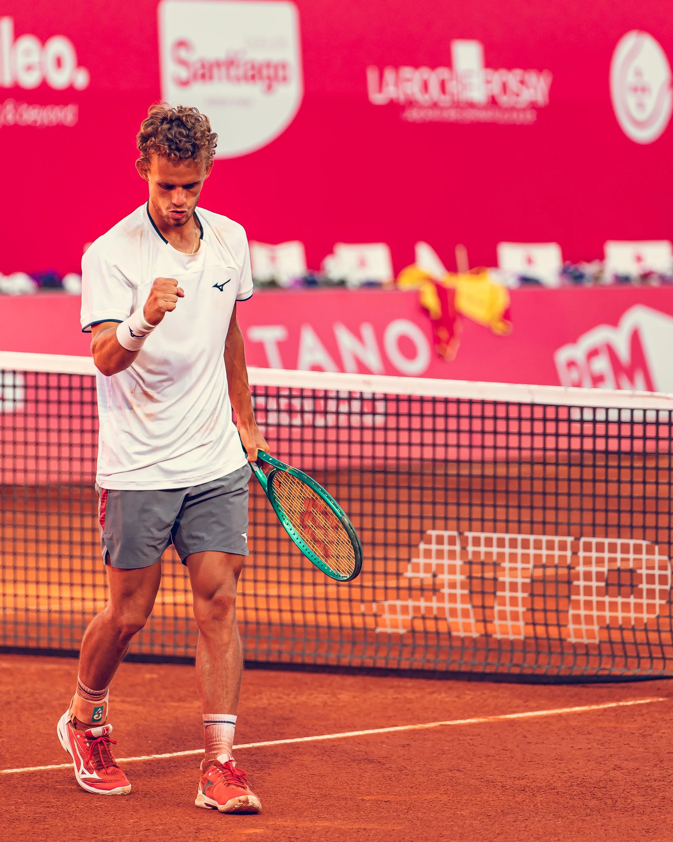 A male tennis player standing on a clay court, wearing a white shirt, gray shorts, and red tennis shoes, holding a tennis racket with a clenched fist.
