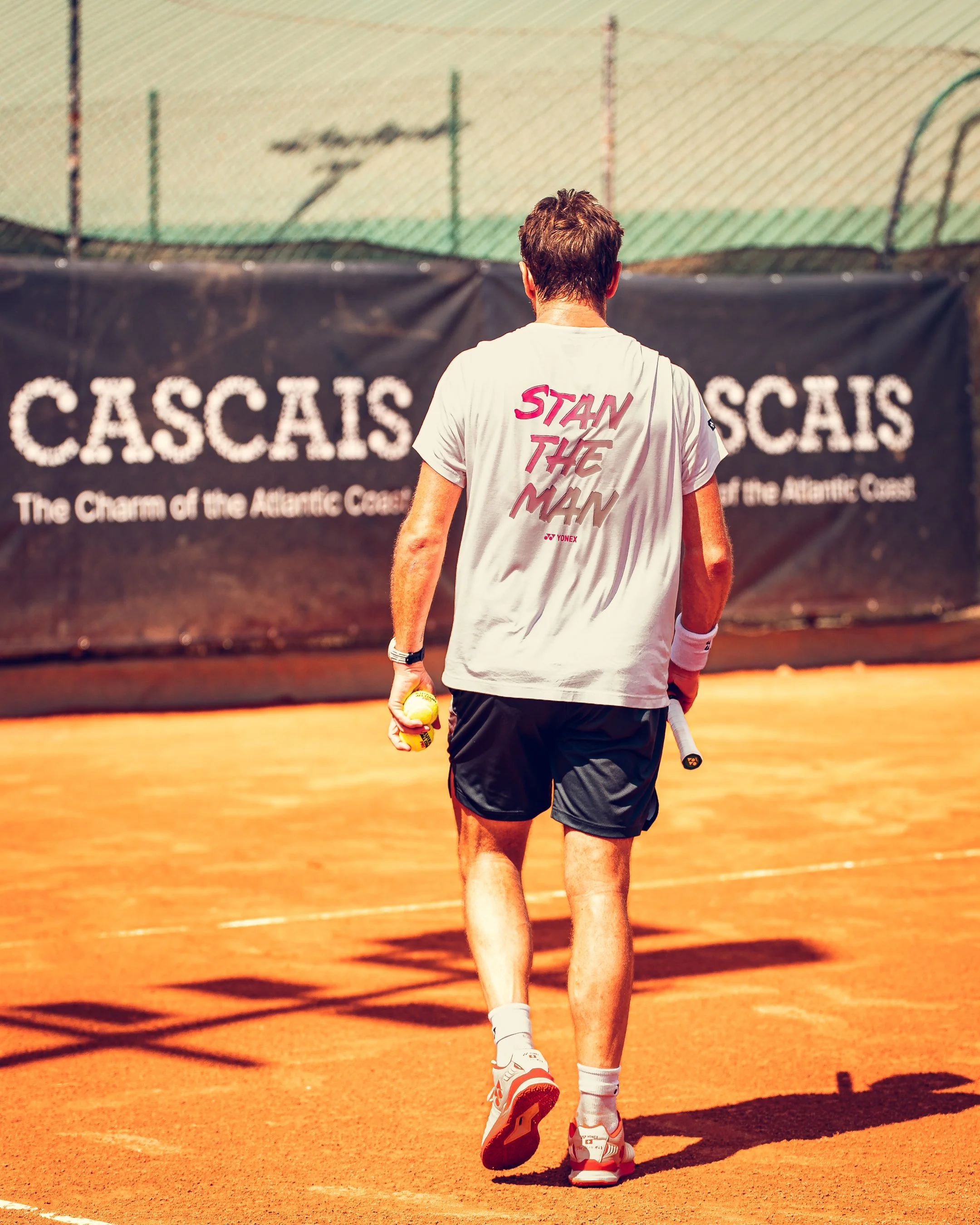 A man walking on a clay tennis court holding a tennis ball in his left hand and a tennis racket in his right hand, with his back facing the camera. He is wearing a gray T-shirt with pink and red text, black shorts, white socks, and white tennis shoes