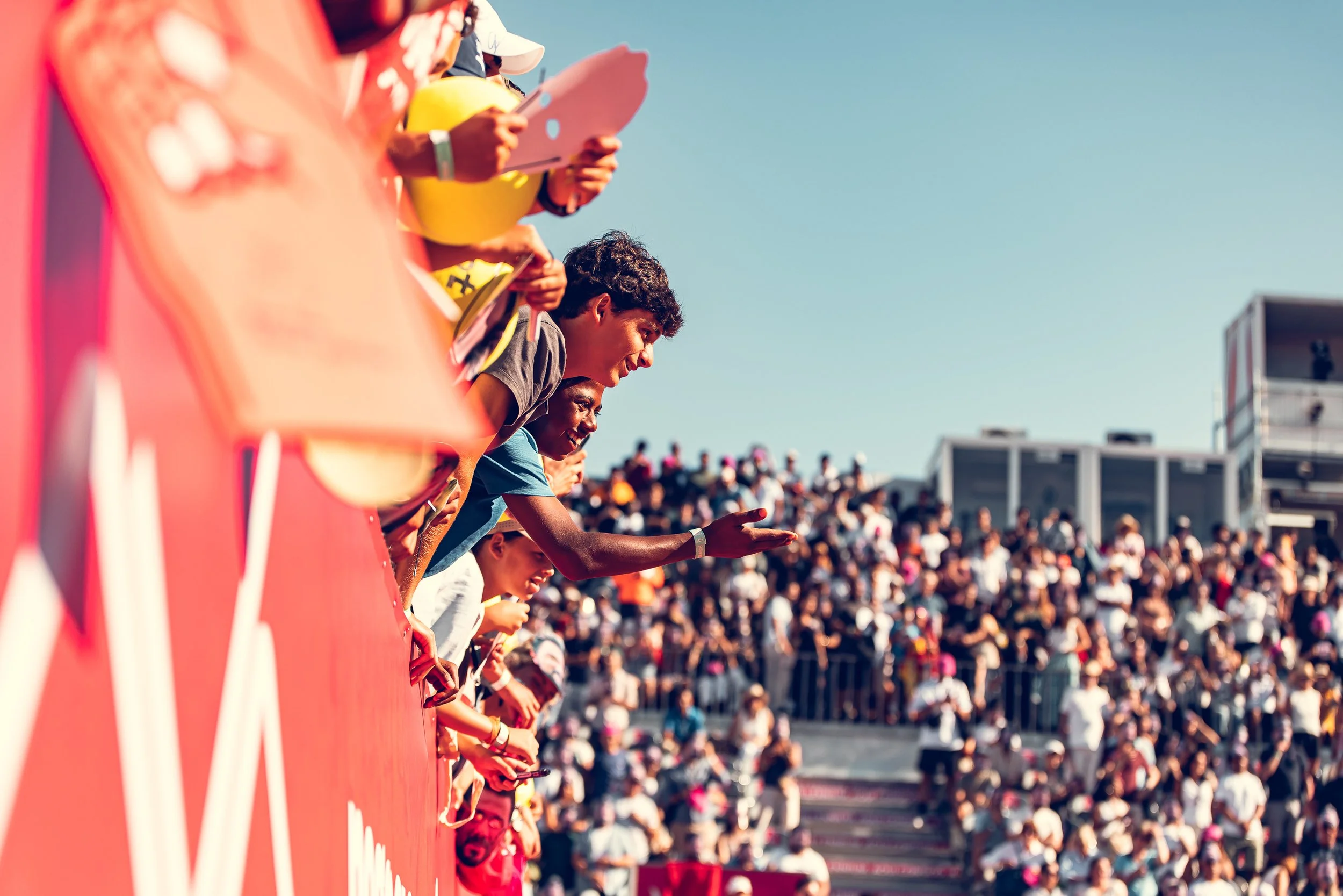 People leaning over a red barrier at a racetrack, looking on and taking photos, with a large crowd and grandstand in the background.