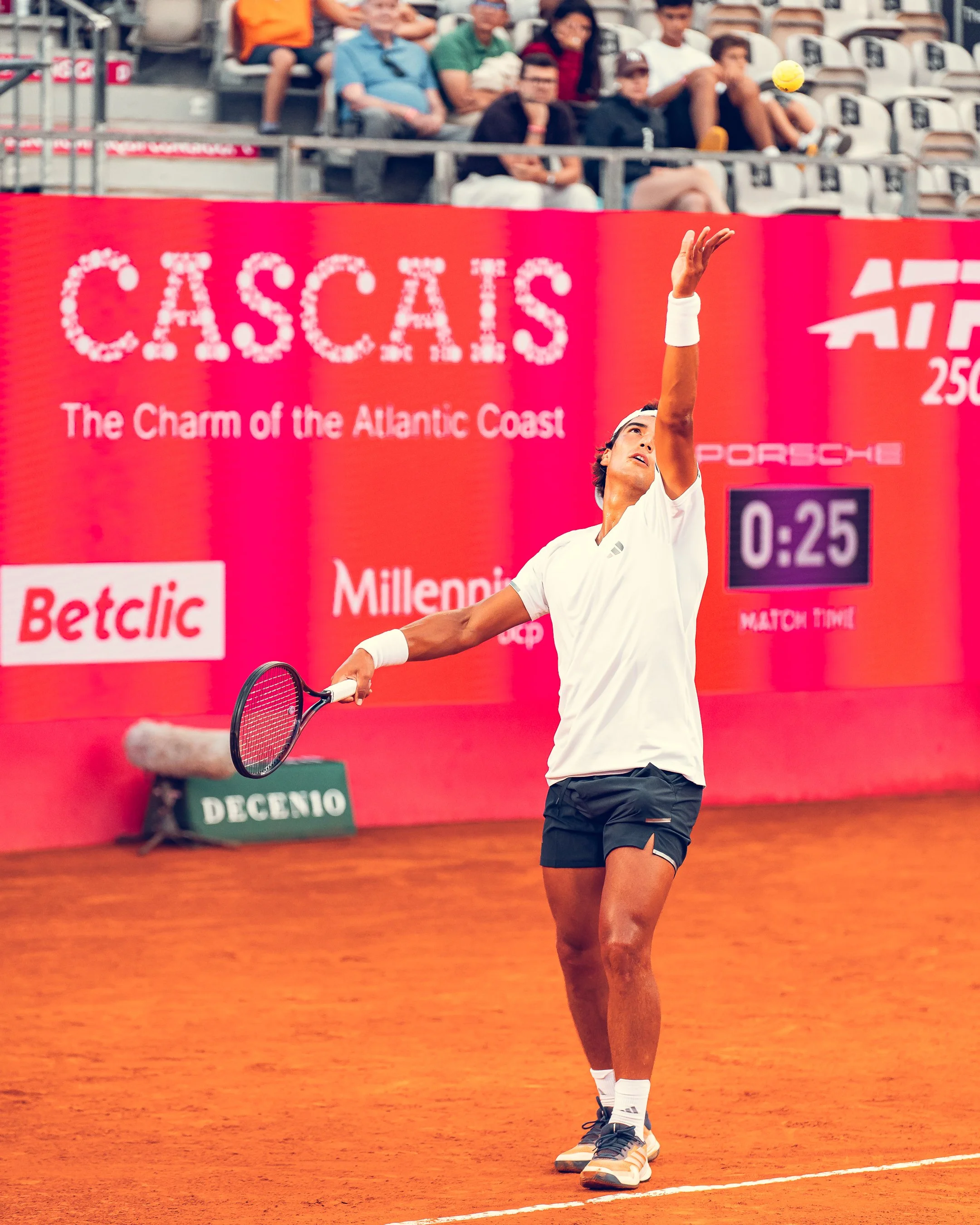 A male tennis player with a white headband, white shirt, black shorts, and tennis shoes, reaching up to hit a yellow tennis ball on a clay court. Spectators are seated in the background, and a red advertising board displaying 'Casca' and other text s