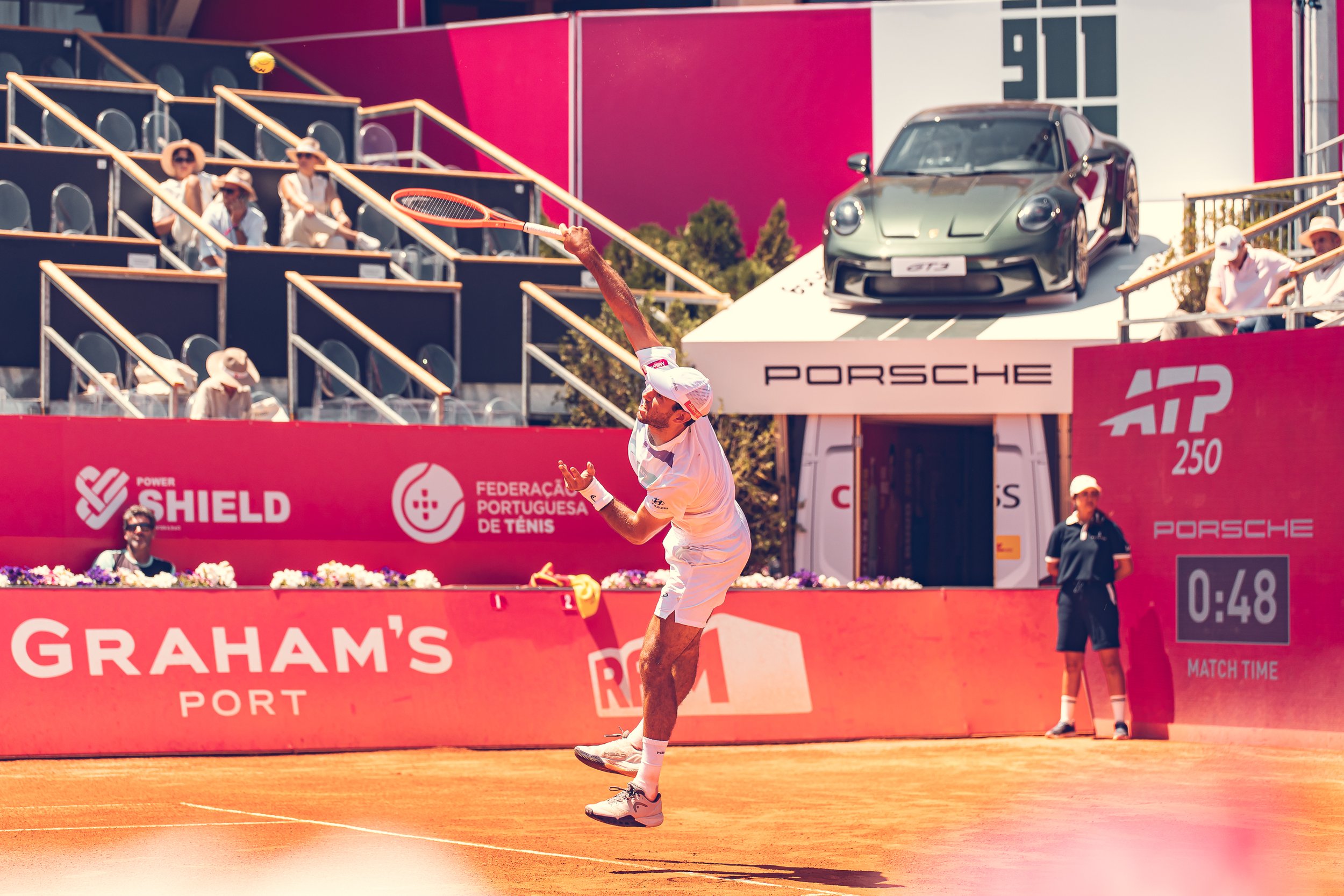 A tennis player wearing a white shirt, shorts, and a cap, hitting a tennis ball with a racket on a red clay court during a match. Spectators are seated in the stands, and a black Porsche race car is displayed in the background.