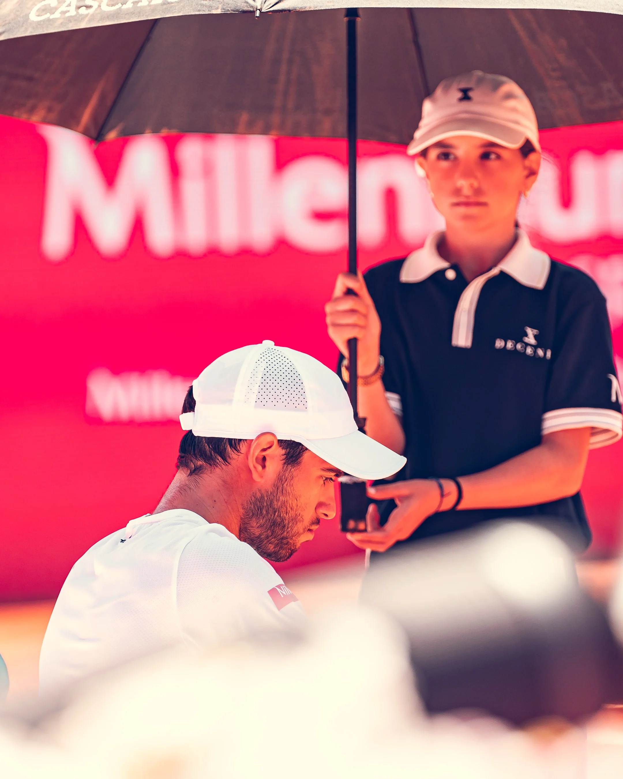 A close-up of a man with a beard and short hair wearing a white cap and white sports shirt, seen from the side. In the background, a woman in a black polo shirt with white collar details and a beige cap is holding an umbrella. The background features