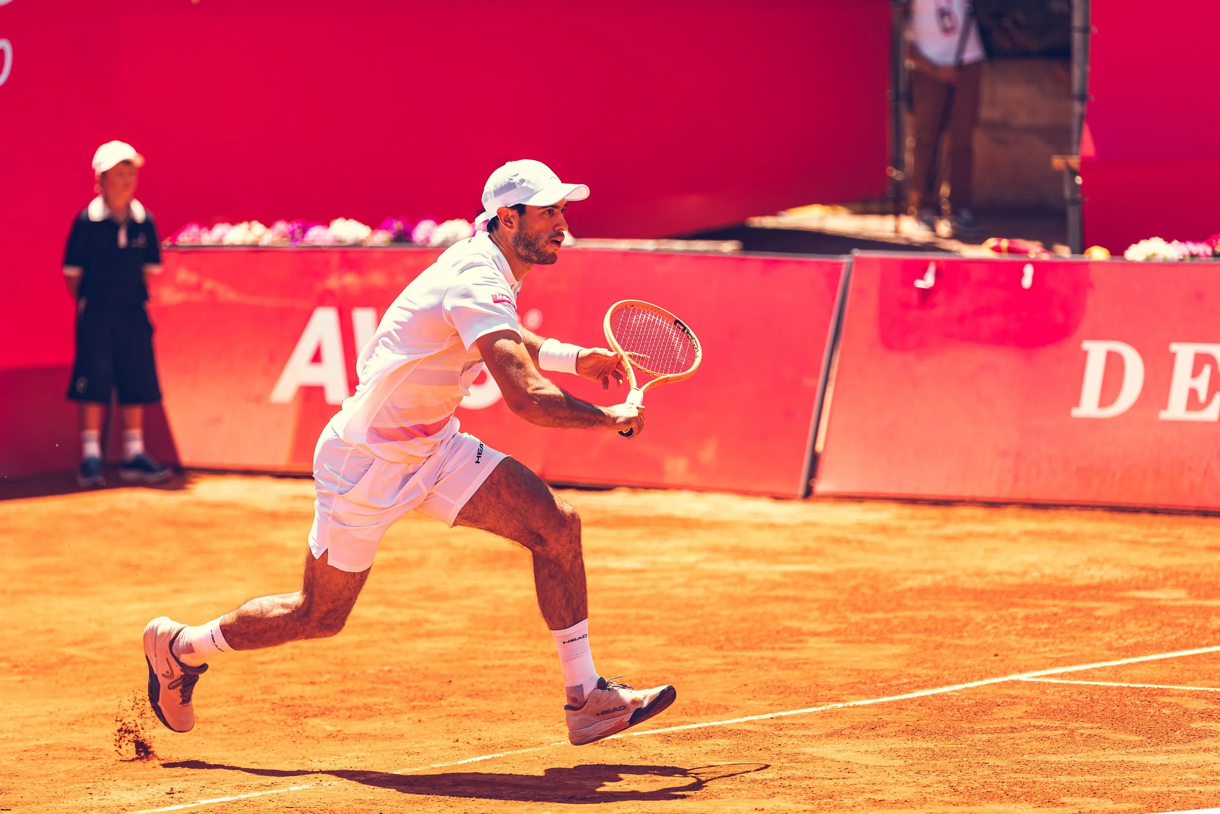 A male tennis player in white sportswear and a cap crouches on a clay court, preparing to return a shot, with a woman in black sportswear and a cap in the background.
