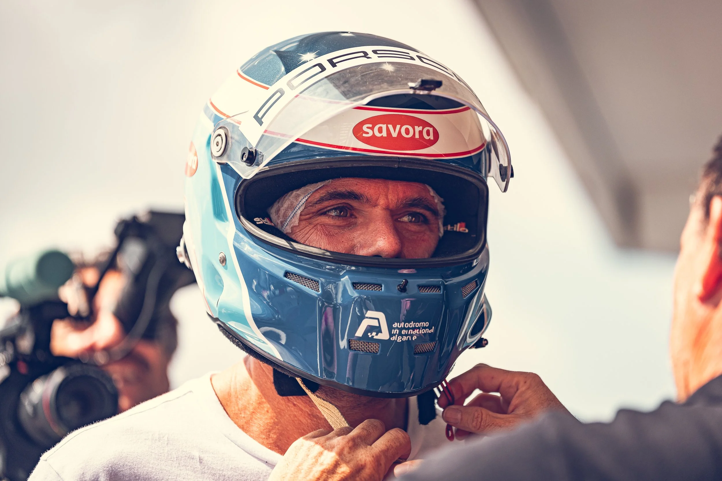 A race car driver is putting on a blue helmet with a clear visor, which has the word 'Porsche' on the top and a 'Savor' sticker on the front. The driver is smiling and looking up, wearing a white shirt, with a person in the background.