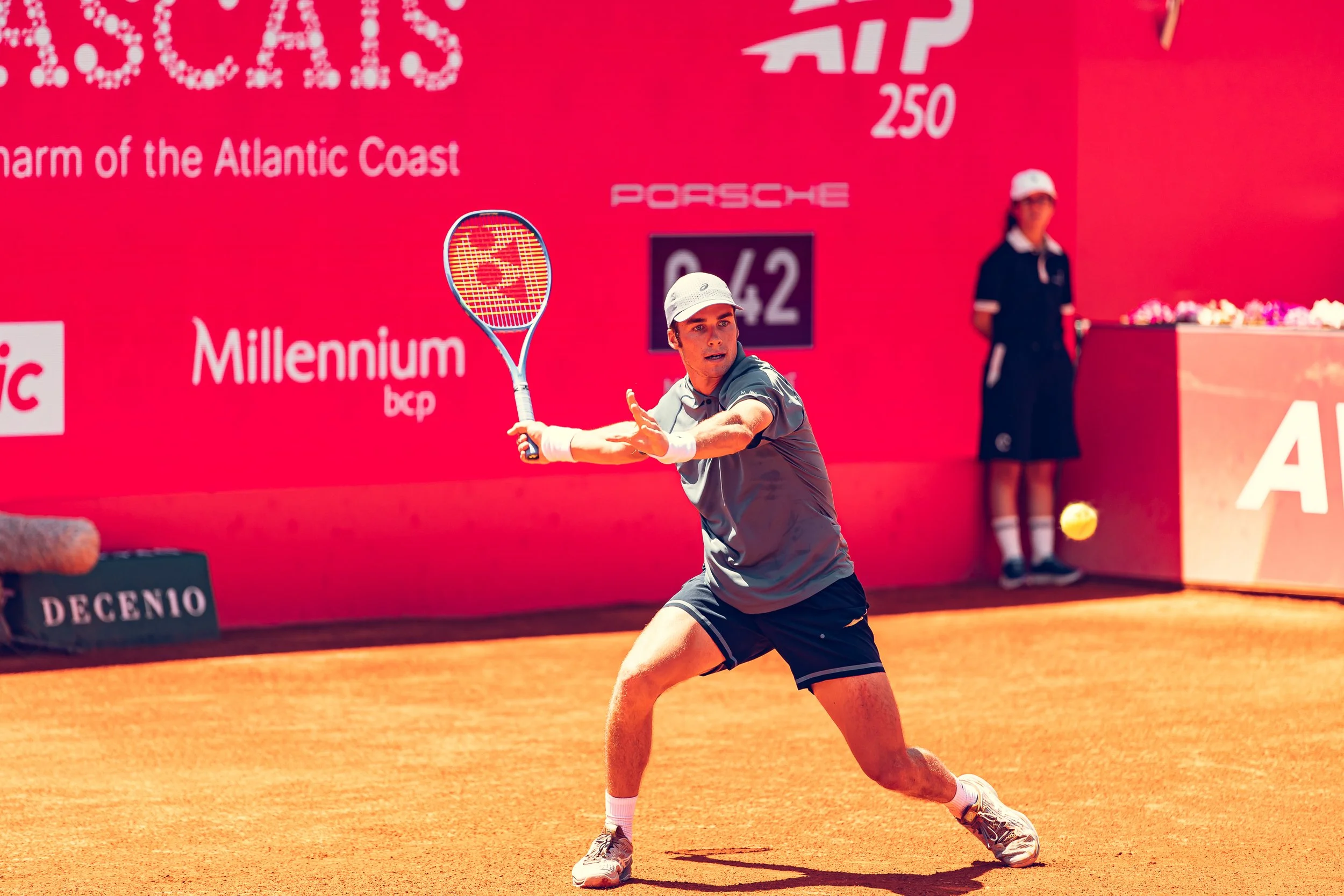 A male tennis player in action on a clay court, preparing to hit a yellow tennis ball. He is wearing a gray shirt, black shorts, a white cap, and tennis shoes. In the background, there is a red wall with sponsor logos and a woman standing near the wa