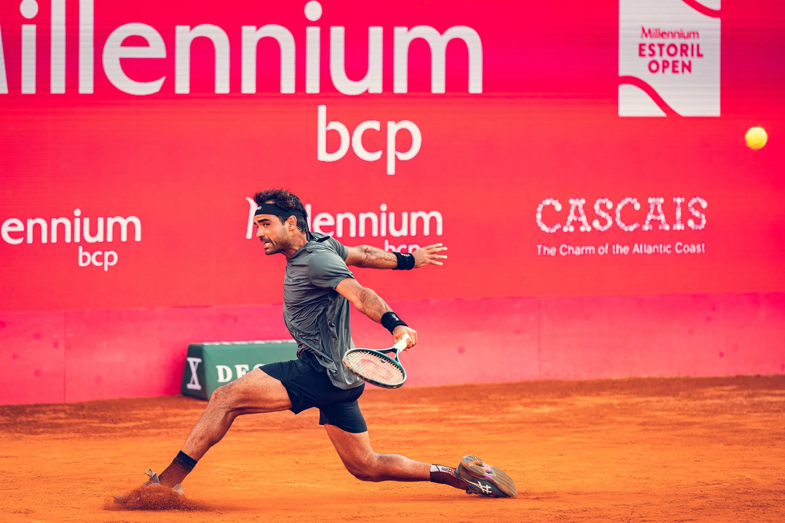 A male tennis player in mid-swing on a clay court, with a bright pink background featuring large white text and logos, including 'Millennium' and 'Estoril Open'.