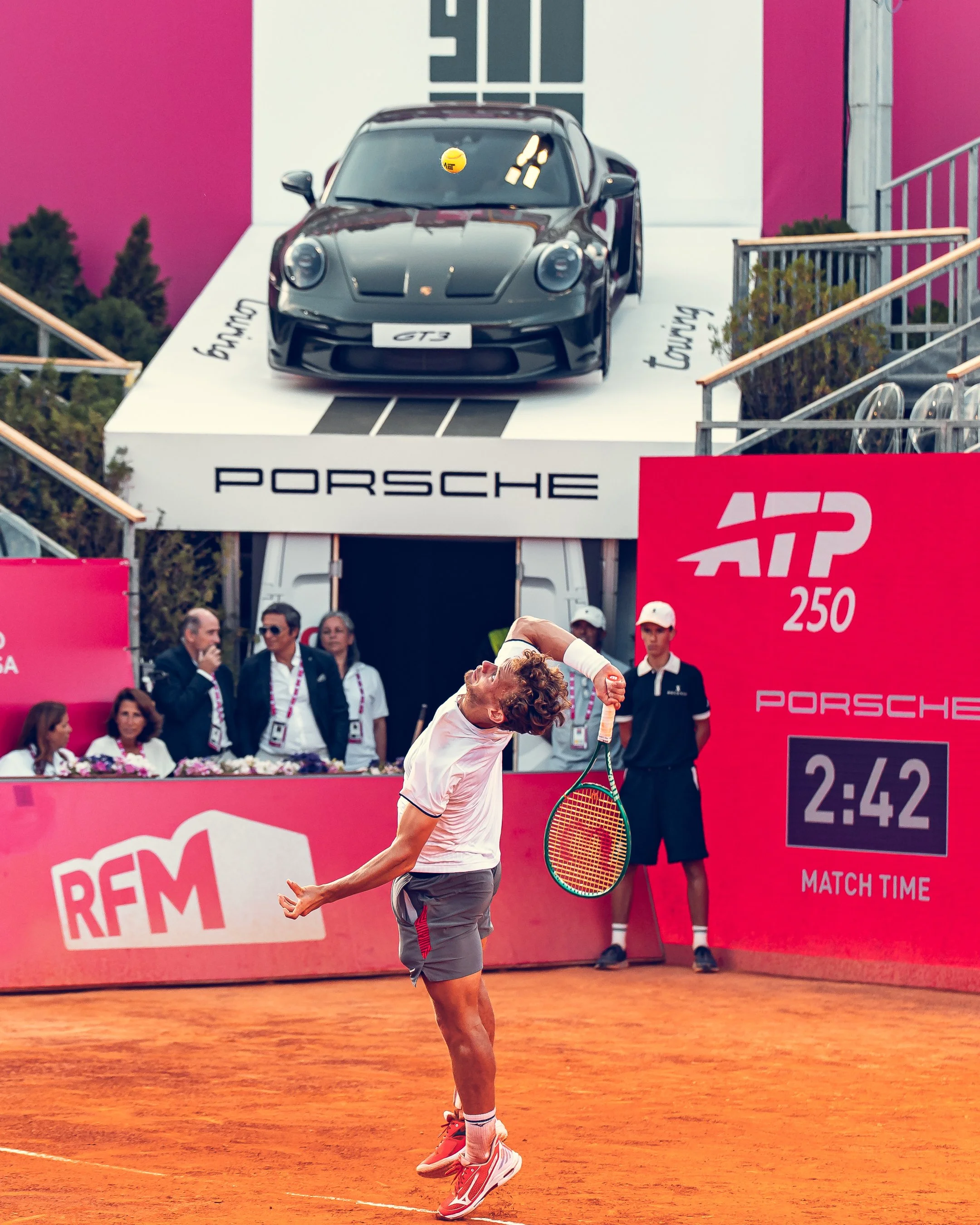 A male tennis player in a gray shorts and white shirt is celebrating on a clay tennis court with a tennis racket in his right hand, and his hair are tousled, with his head tilted back and eyes closed. In the background, there are several people and a