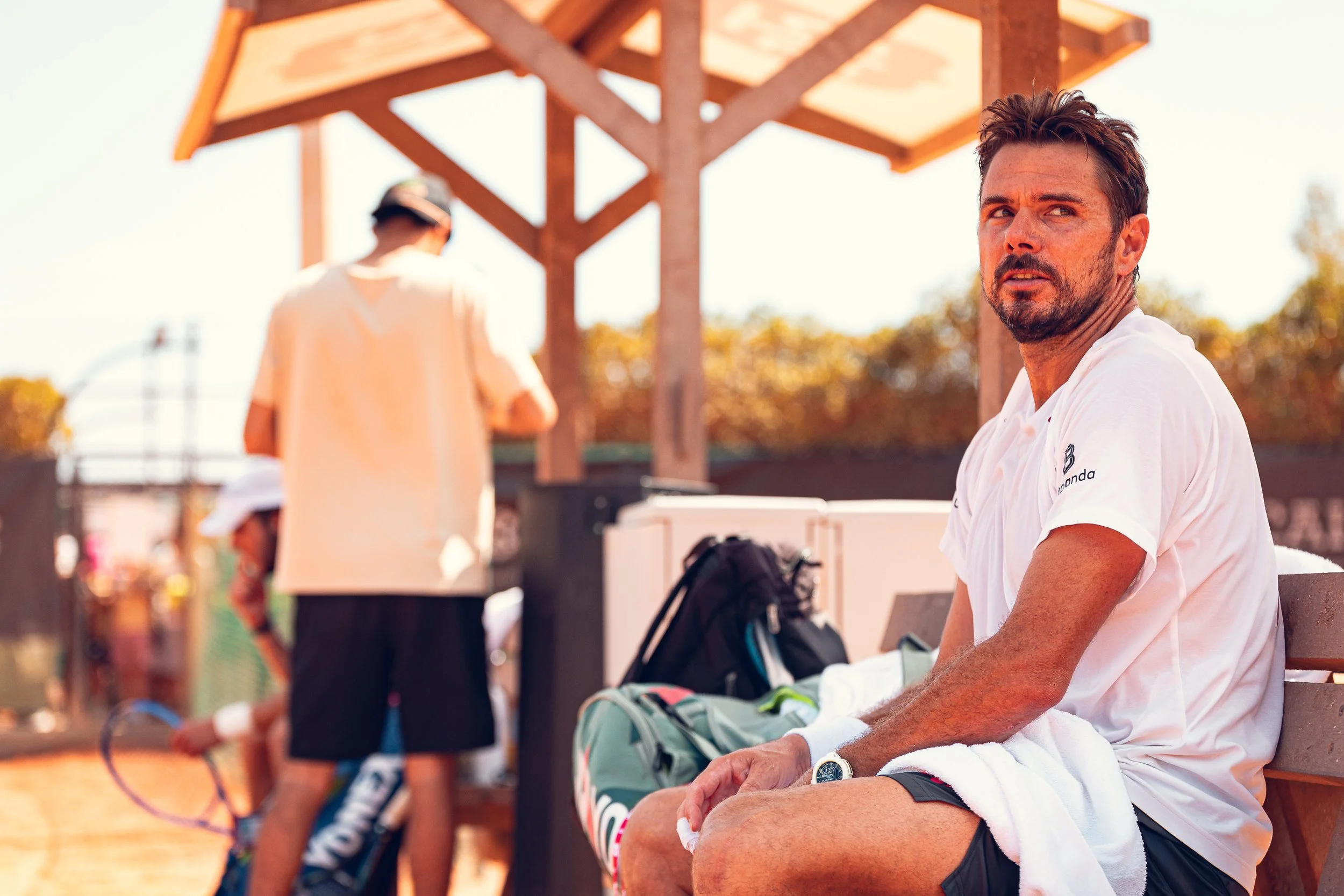 Man with dark hair and beard sitting on a bench, wearing a white shirt, with tennis bags and equipment nearby. Background shows a person wearing a hat and shorts, and a wooden structure.