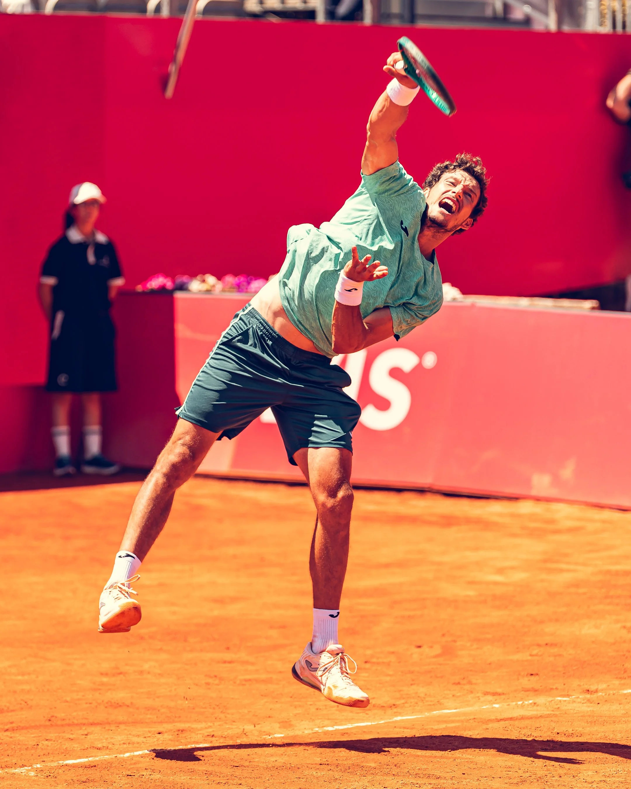 A male tennis player in a green shirt and shorts, with white wristbands, is hitting a tennis ball on a clay court. He is wearing white athletic shoes and appears to be in mid-motion while playing. A line judge or opponent dressed in black stands in t