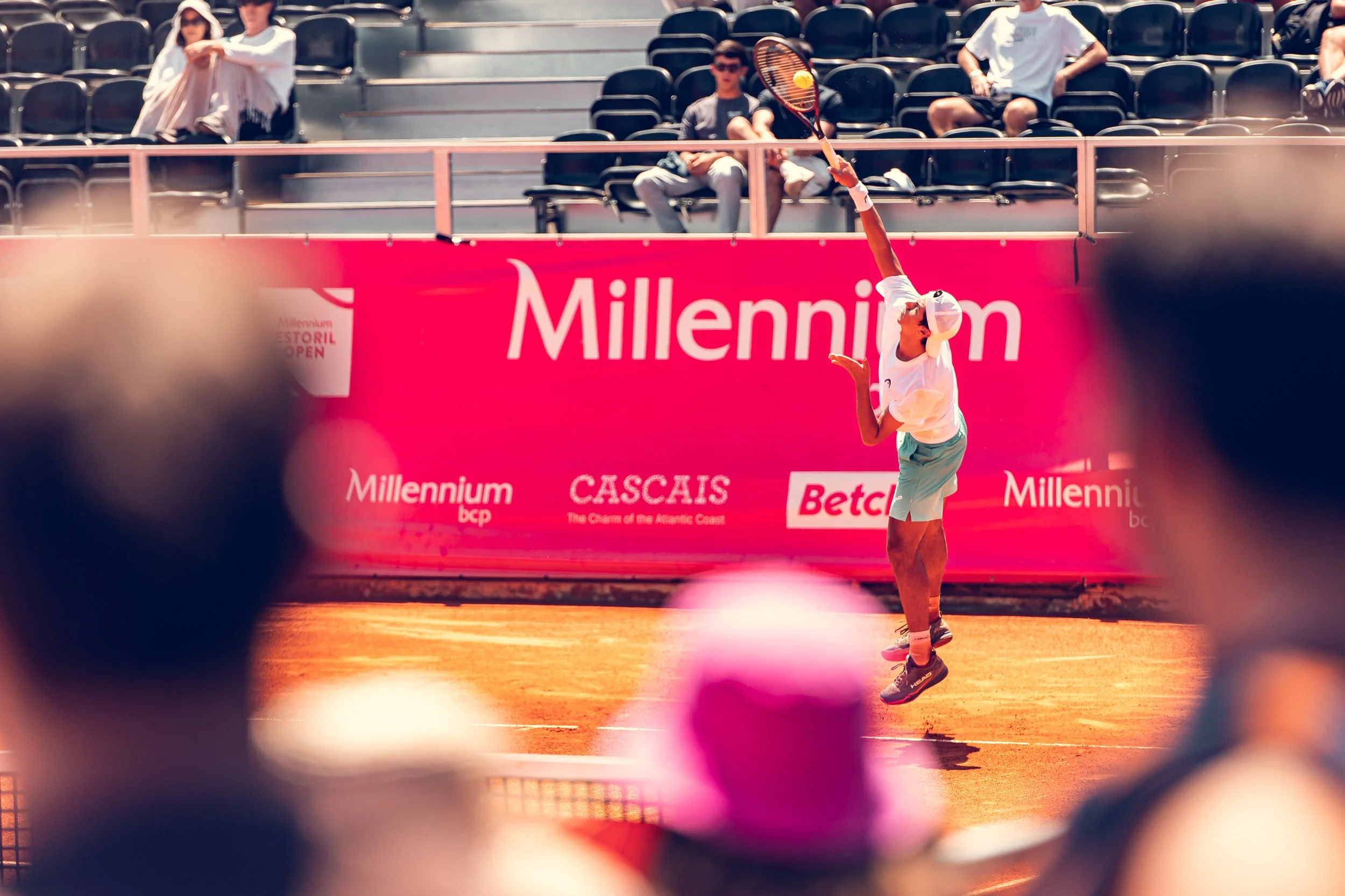 A female tennis player in white athletic clothing and a cap serving the ball on a clay tennis court during a match. Spectators are seated in the background behind a bright pink advertising banner.