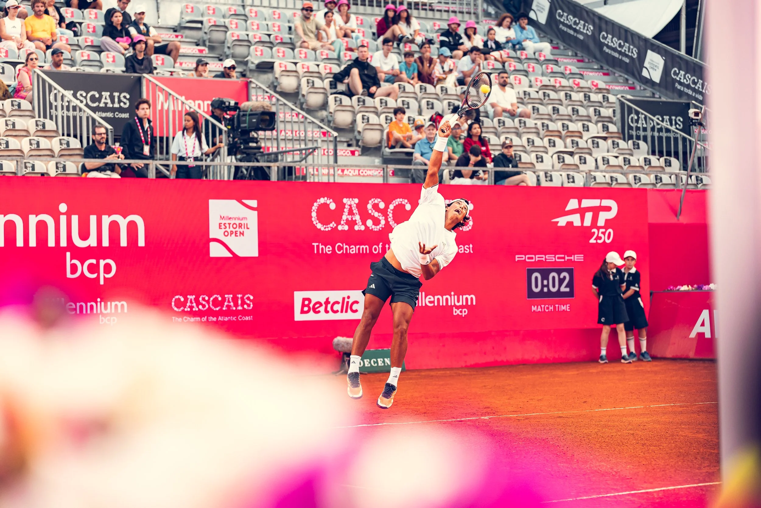 A female tennis player in mid-air hitting a tennis ball with a racket during a match at an outdoor tennis stadium, with spectators seated in the background.