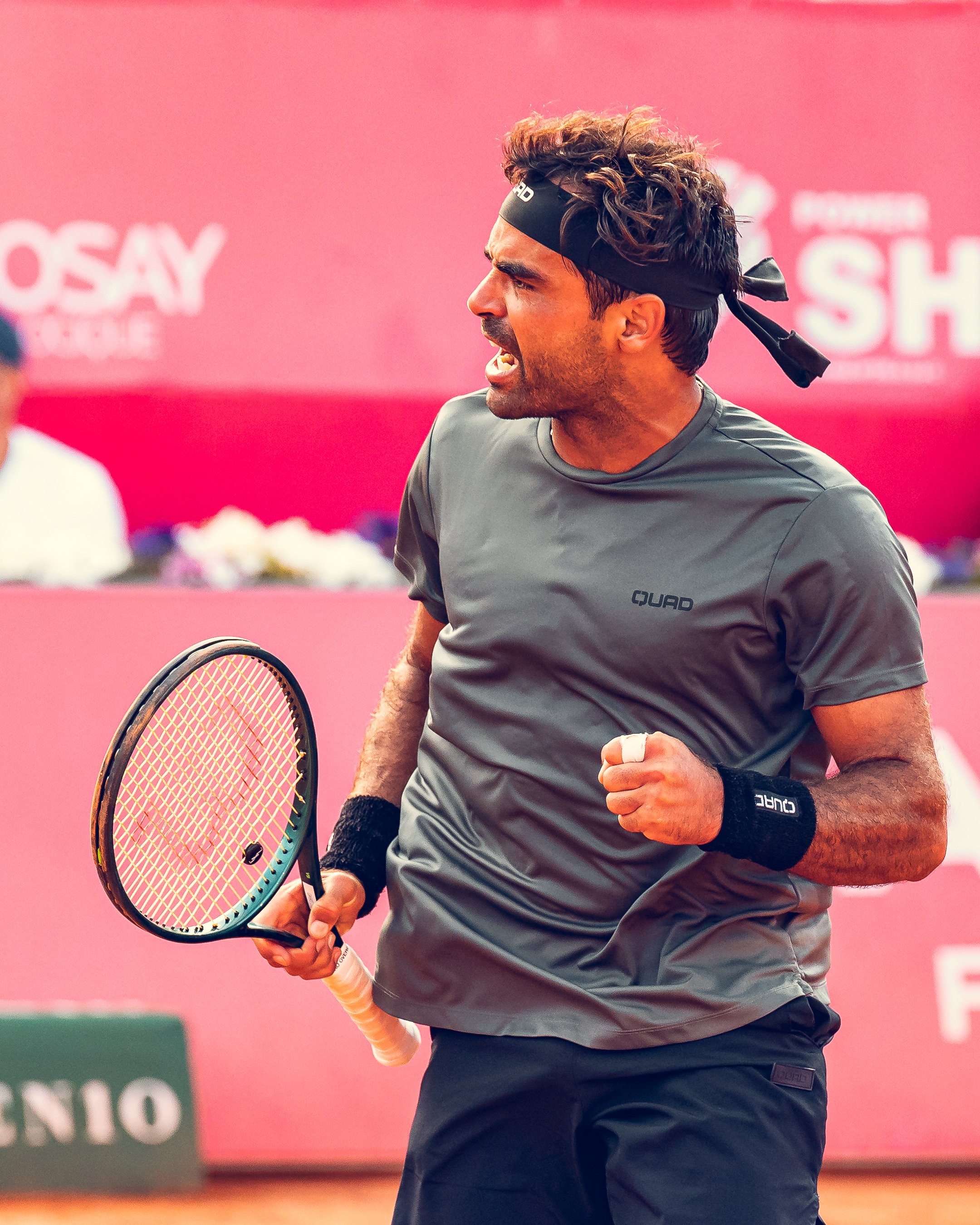 A male tennis player in a gray shirt and black shorts celebrating on a tennis court, holding a tennis racquet in his right hand, with a pink background.
