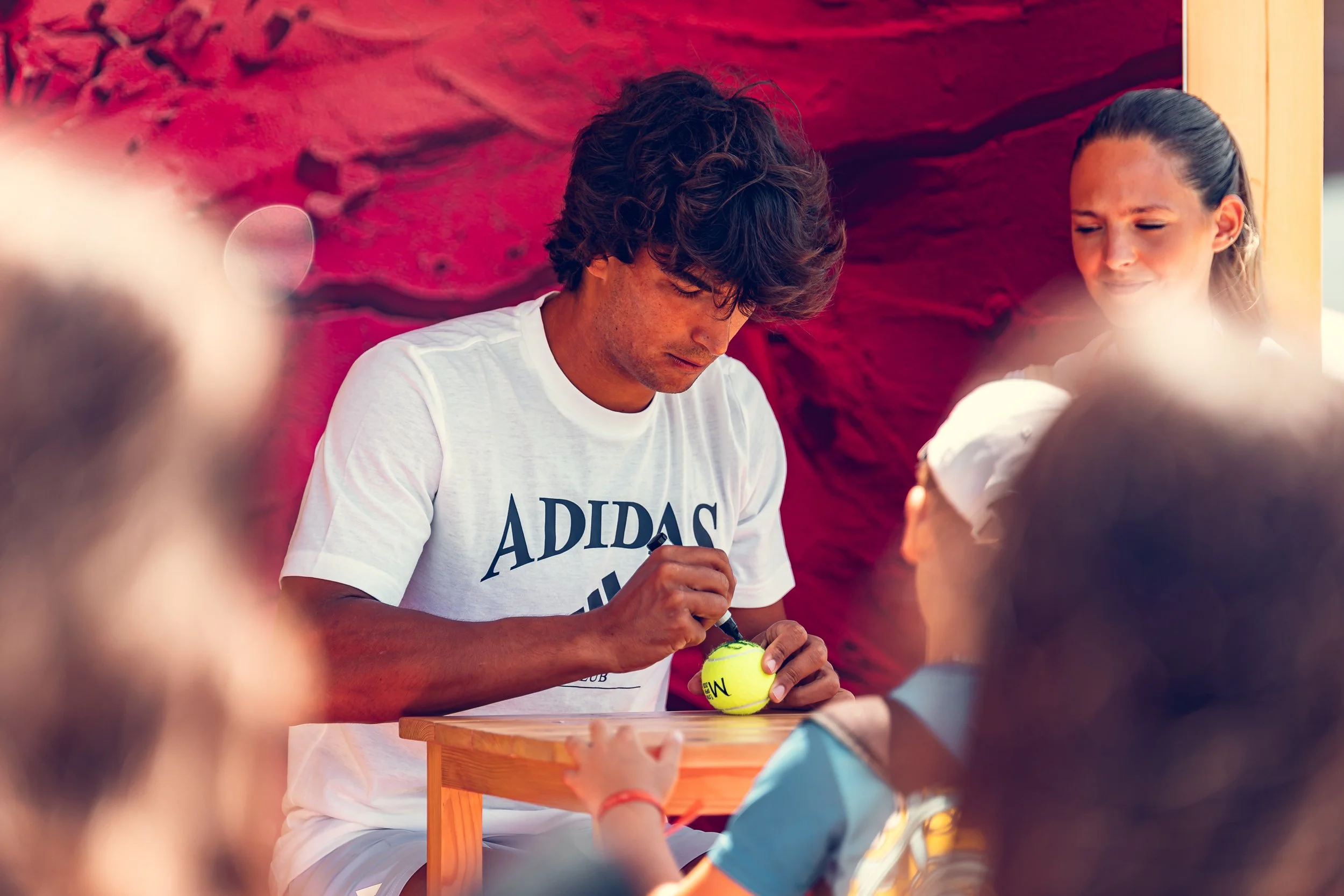 A person with curly hair wearing a white Adidas t-shirt is signing tennis balls at an event, with children and blurred people in the foreground against a red rocky background.