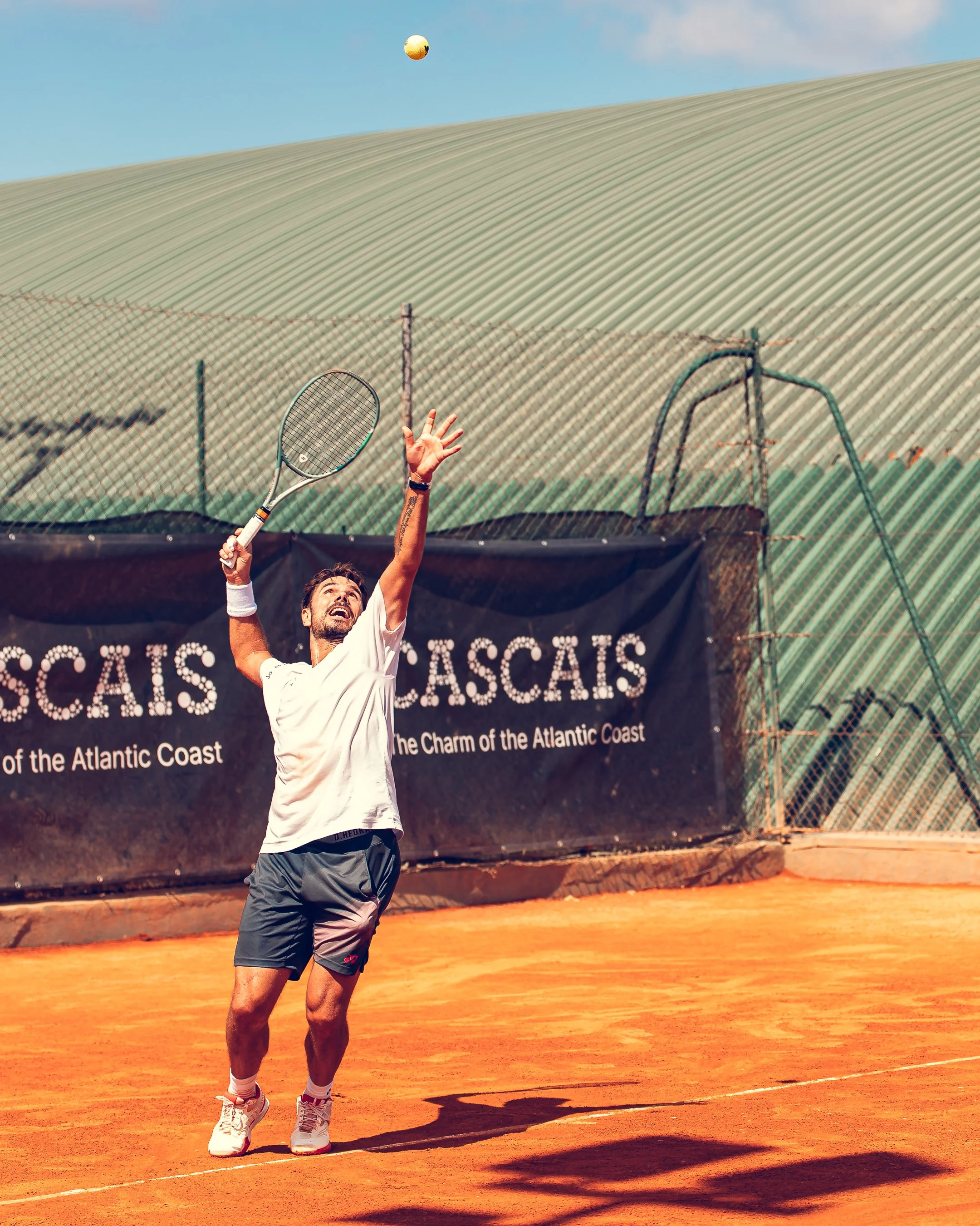 Male tennis player on red clay court, reaching to hit tennis ball with racket, wearing white shirt, gray shorts, and white sneakers, with tennis court fence and green landscape in background.