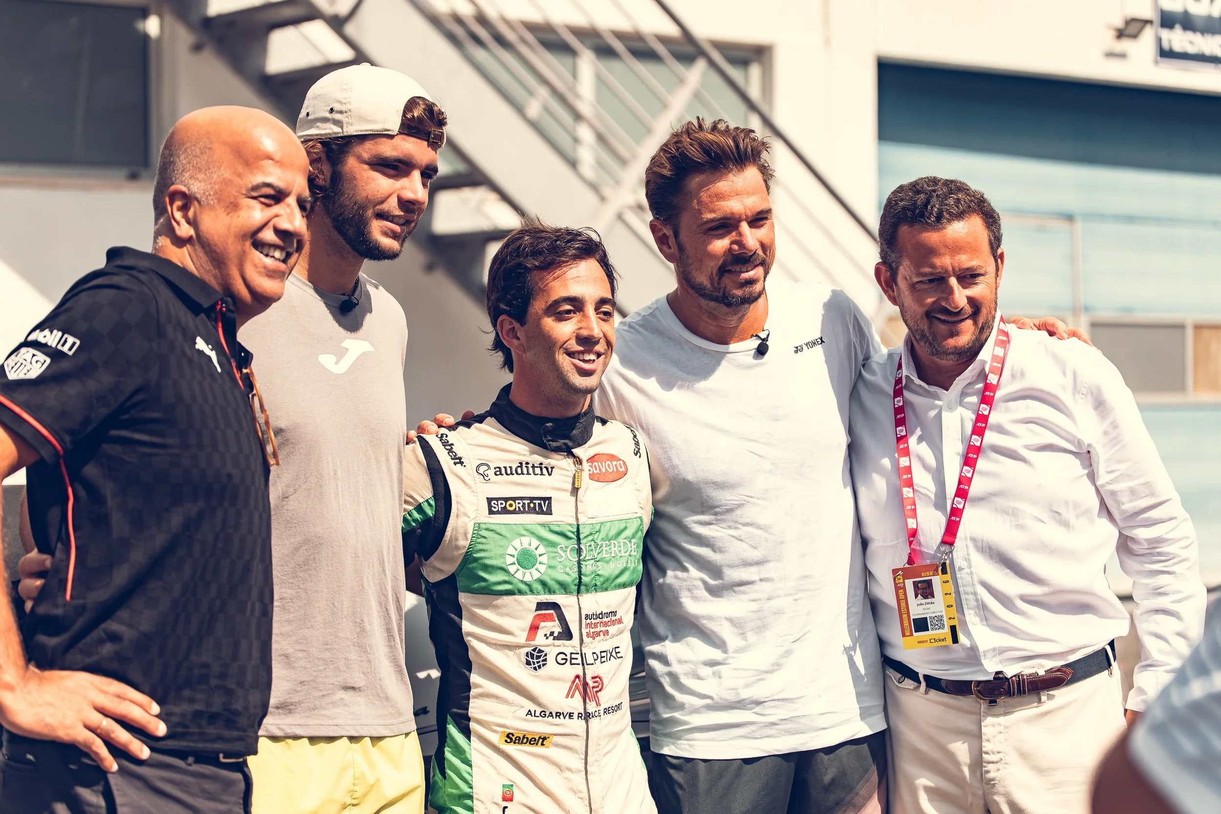 Group of six men standing together at a race event, smiling and posing for a photo. One man is dressed in a racing suit, while the others are in casual or official attire. They are outdoors, with a building and track visible in the background.