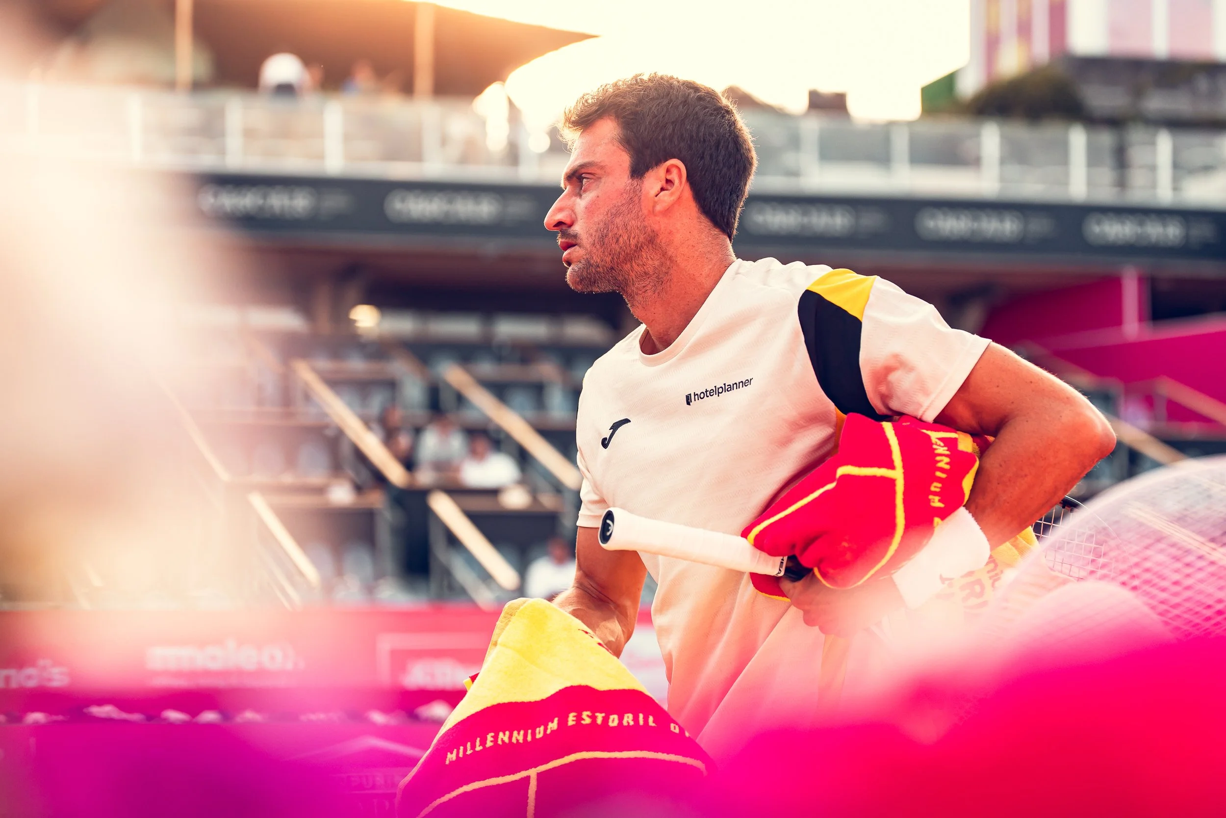 A male tennis player with short dark hair and beard, wearing a white sports shirt with the logo 'hotelplanner' and a yellow and red towel draped over his arm, stands on an outdoor court during a match at sunset.