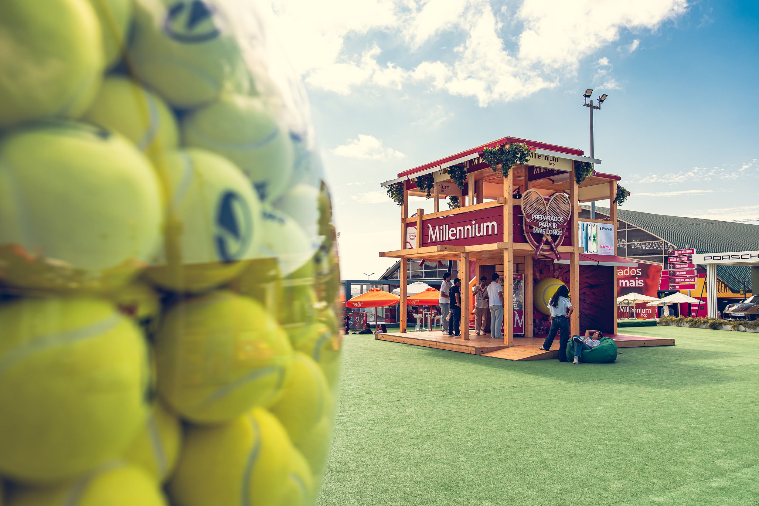 A cluster of yellow tennis balls in the foreground on the left side. In the background, there is a wooden pavilion with signs displaying the word 'Millennium' and tennis-related imagery. Several people are gathered near the pavilion, and the sky is m