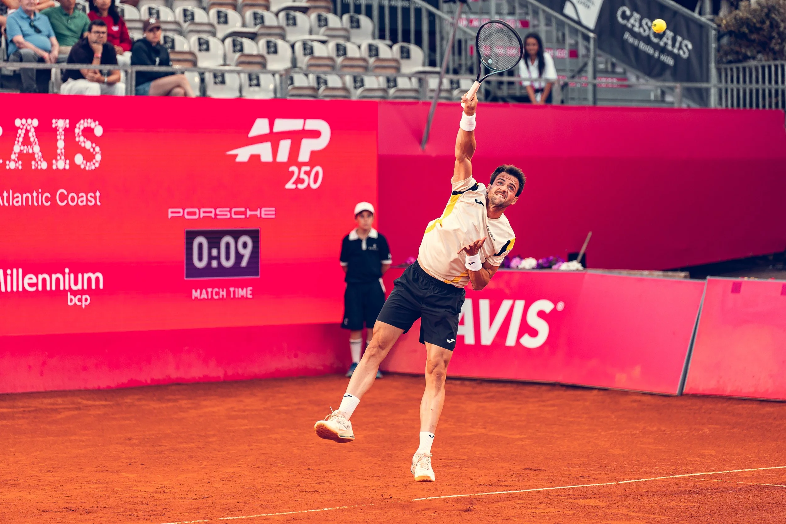 A male tennis player is throwing his racket into the air on a clay court during a match, with spectators watching in the background.