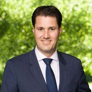 Man in zakelijke kleding met een grijns, groene boombladeren op de achtergrond.