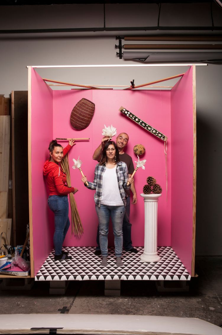 Image of three people standing on a three walled box painted bright pink with miscellaneous objects on the walls, in their hands, and on a column places within the box.