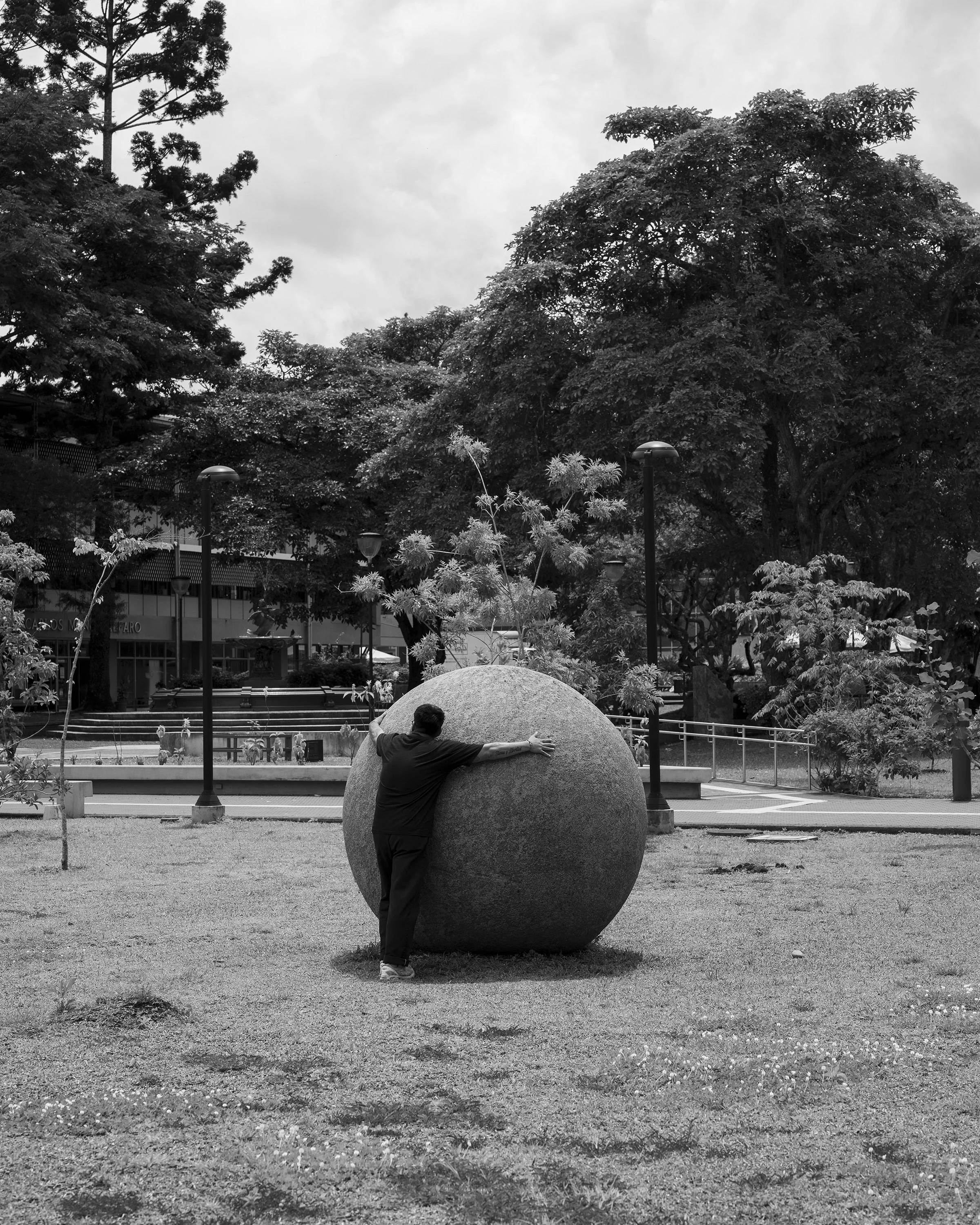 Black and white photo of a man hugging a giant concrete ball in a park, by William Camargo