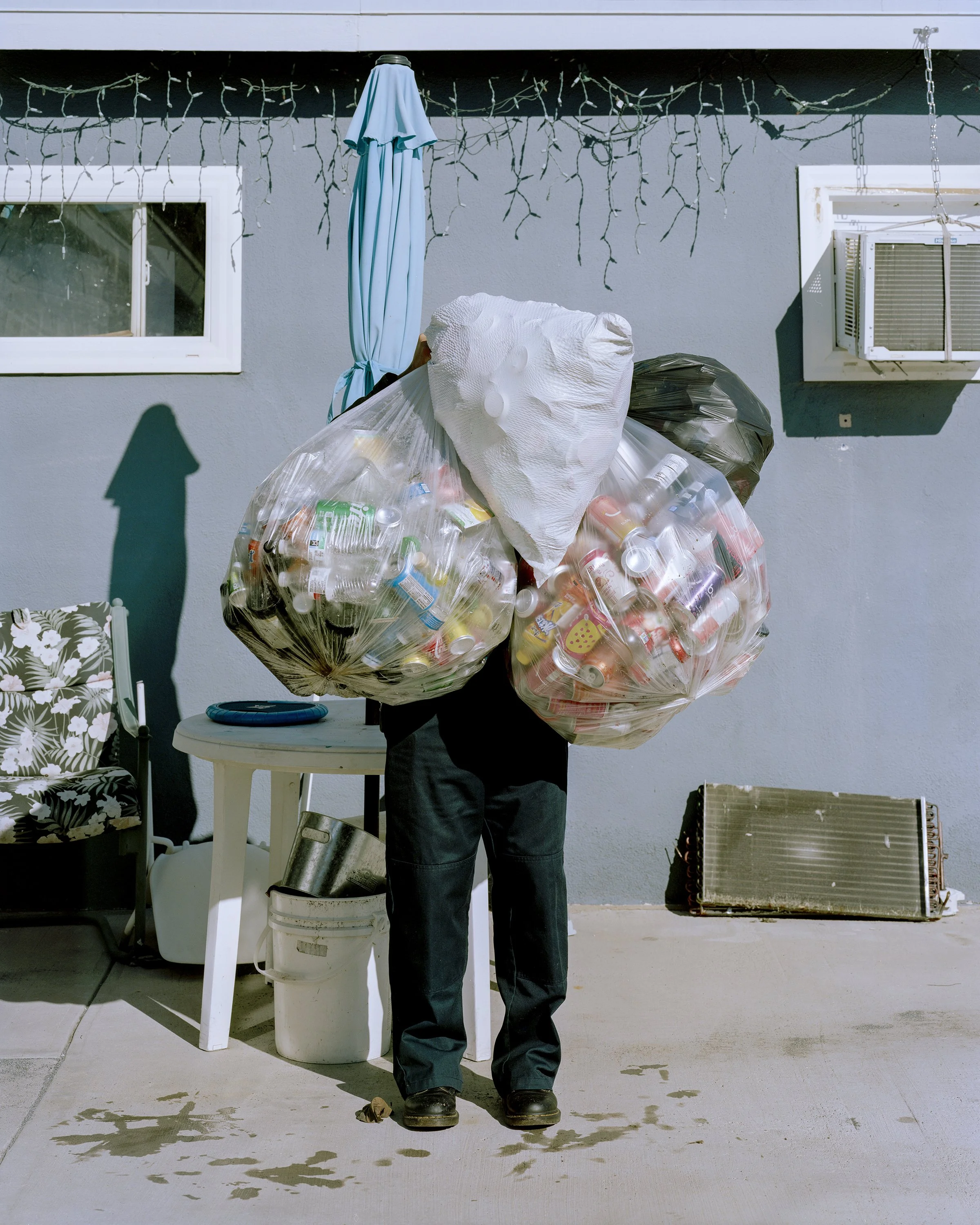 Color photograph of a person carrying four large trash bags filled with cans and bottles obscuring their face, by William Camargo